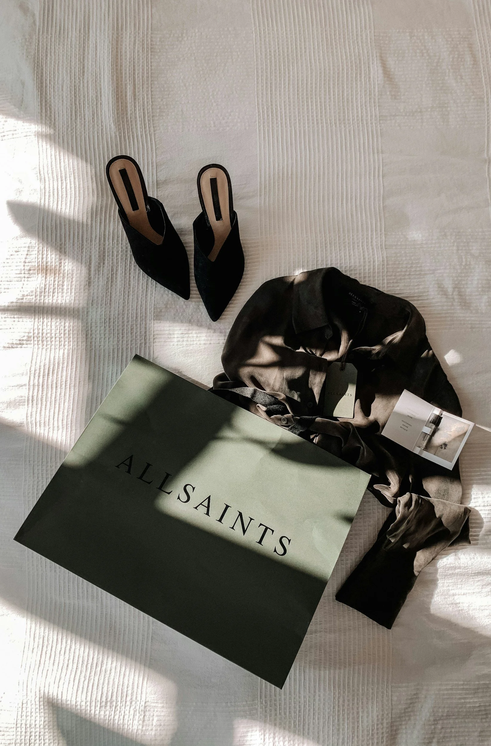 Zapatos negros de tacón alto, un saco y una caja de tienda de calzado llamada 'ALLSAINTS' en la cama.