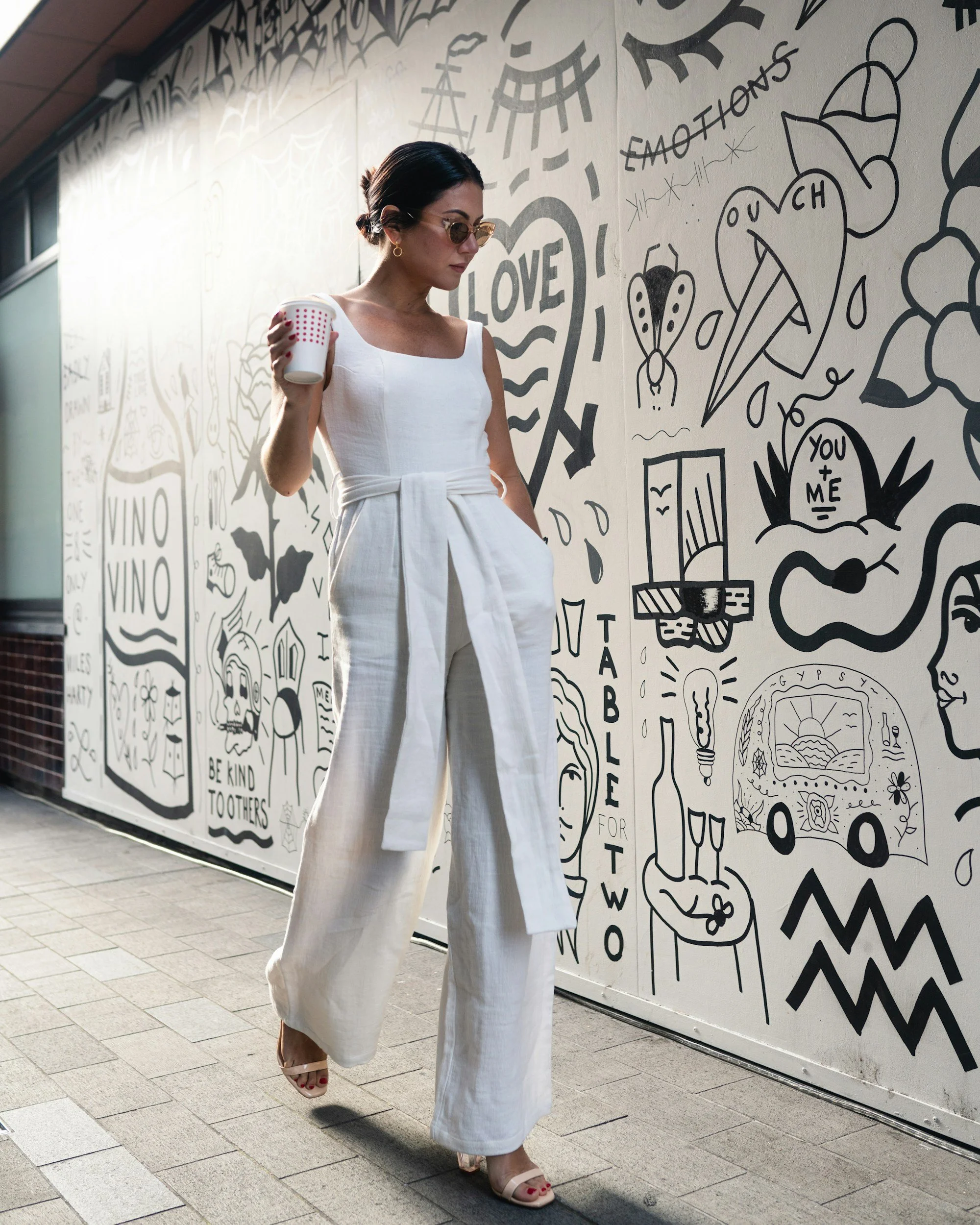 Mujer con vestido blanco y gafas de sol caminando junto a una pared con dibujos en blanco y negro que incluyen corazones, palabras como I LOVE y YOU + ME, y ilustraciones como mariposas y escenas