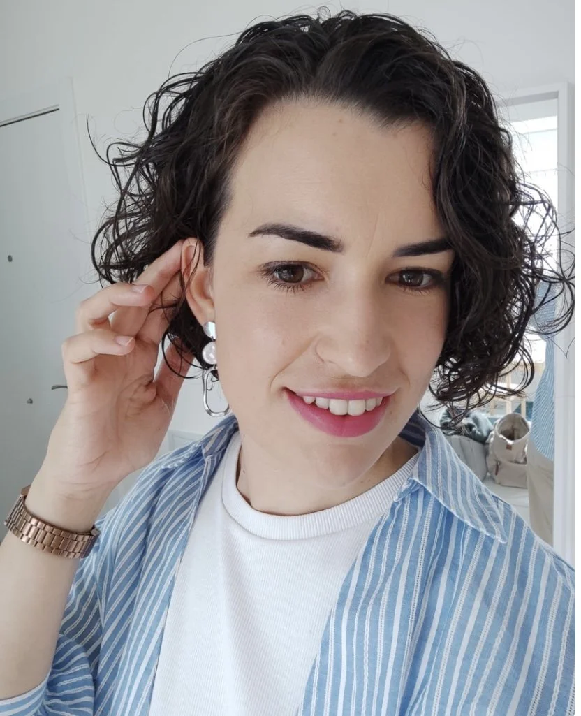 Retrato de una mujer con cabello rizado y oscuro, usando pendientes grandes y un reloj dorado, sonriendo en un ambiente interior con ventanas al fondo.