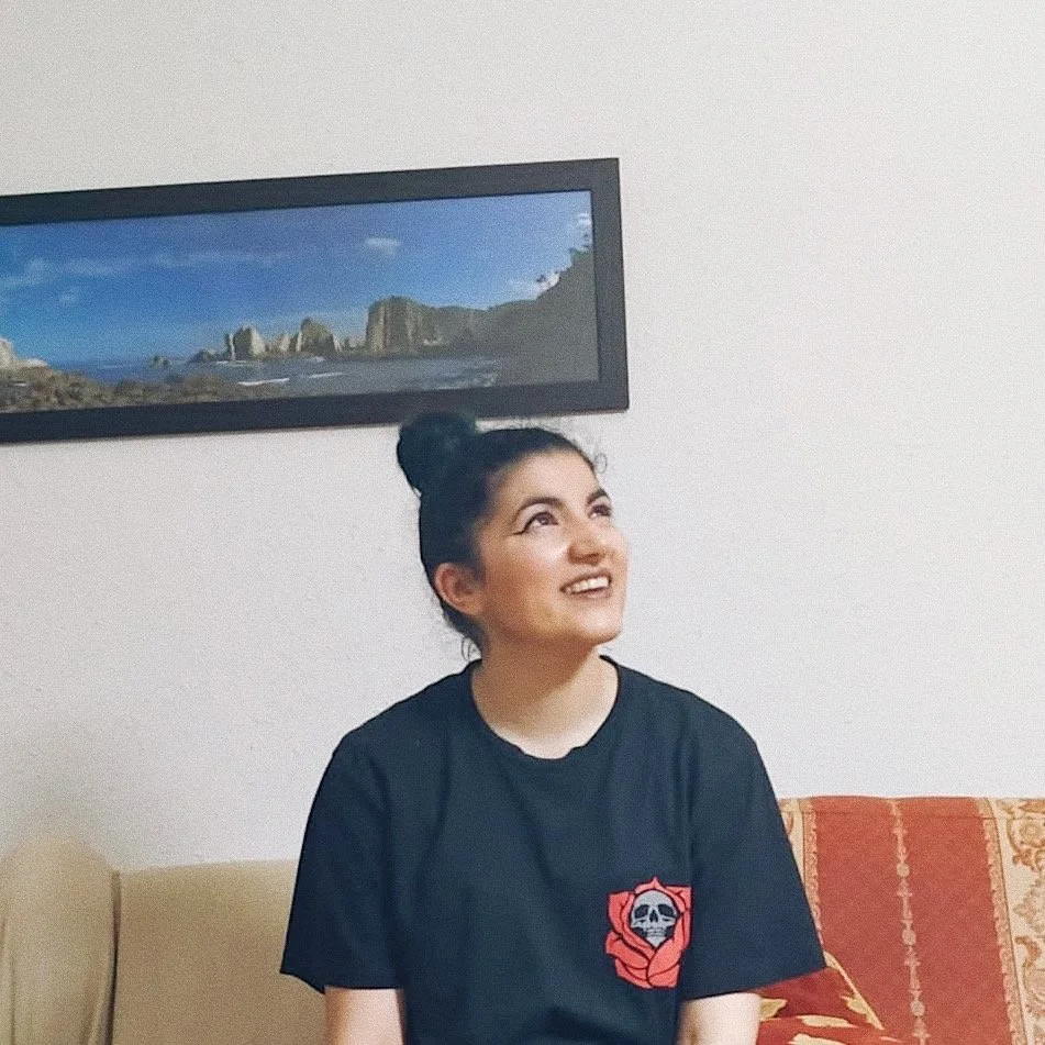 Mujer joven con peinado en moño sentado en un sofá, sonriendo y mirando hacia arriba, en una sala con cuadro de paisajes en la pared.