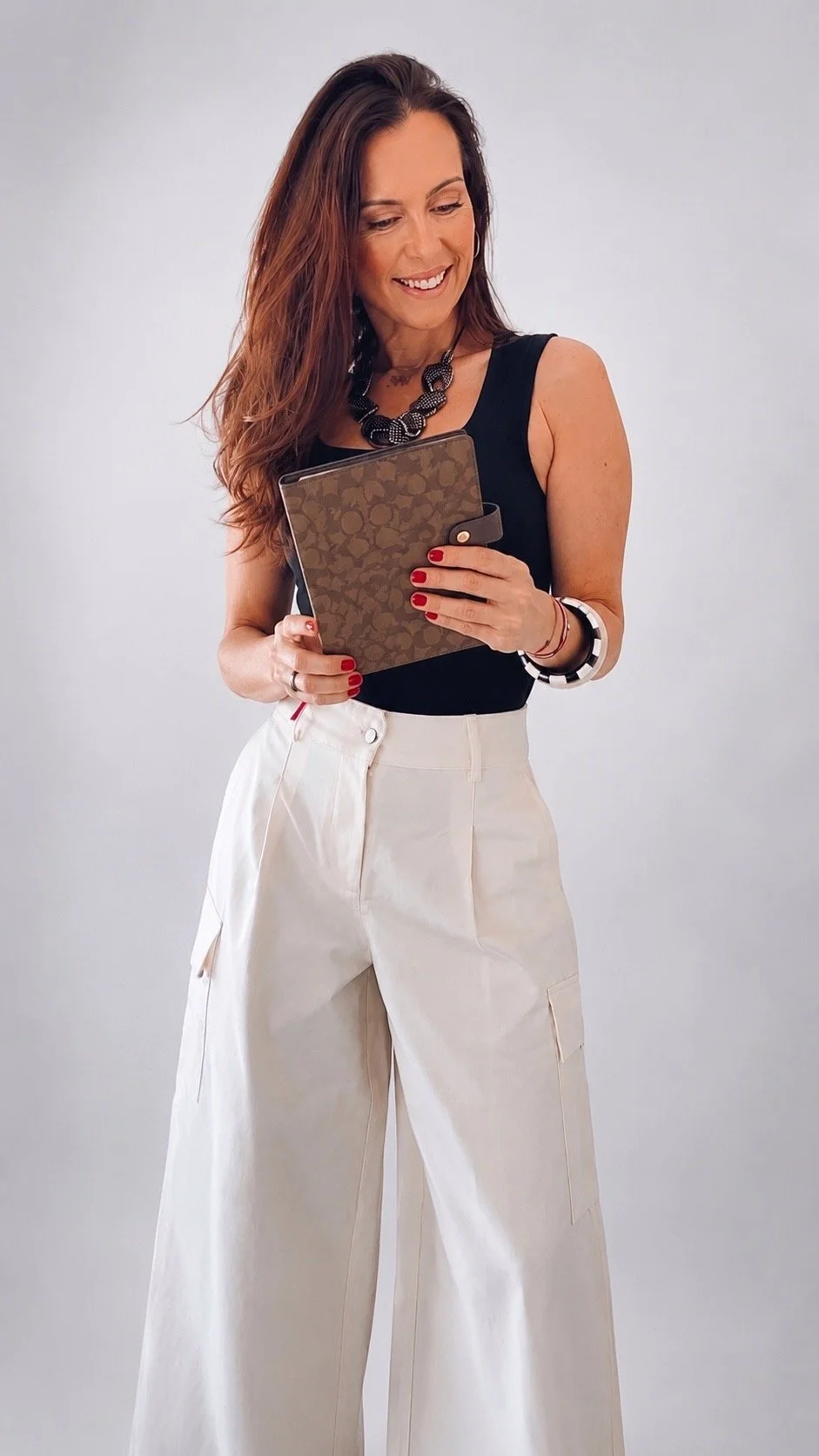 Mujer con cabello castaño oscuro, usando blusa negra sin mangas y pantalones beige anchos, sosteniendo una carpeta o librito, con accesorios como collar negro, pulsera blanca y rojo, y sonrisa mientras mira el contenido de la carpeta.