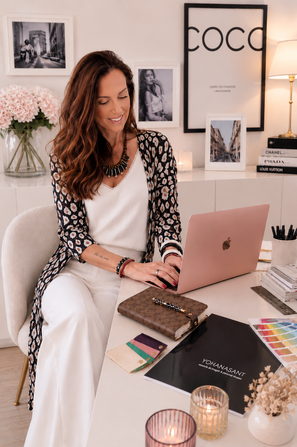 Mujer sonriente trabajando en una oficina moderna, rodeada de libros de marca de lujo, fotos enmarcadas y decoración floral.