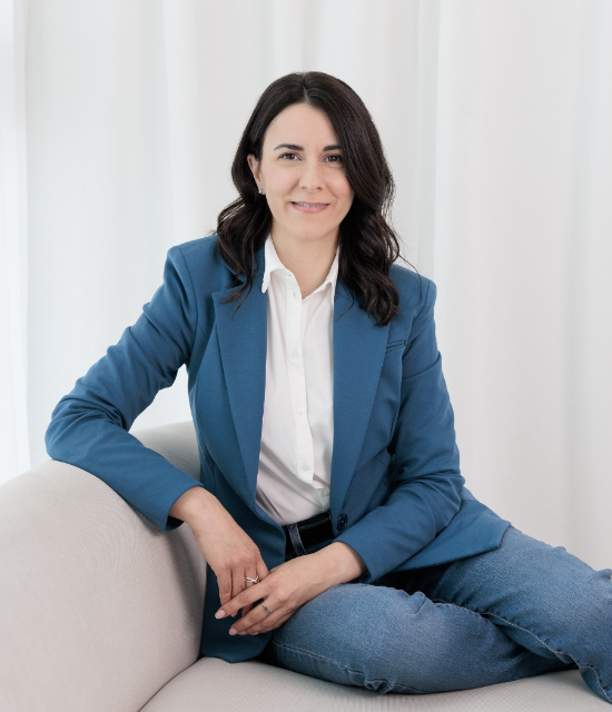 Mujer con blazer azul y jeans sentada en un sofá blanco en un ambiente interior con cortinas blancas.