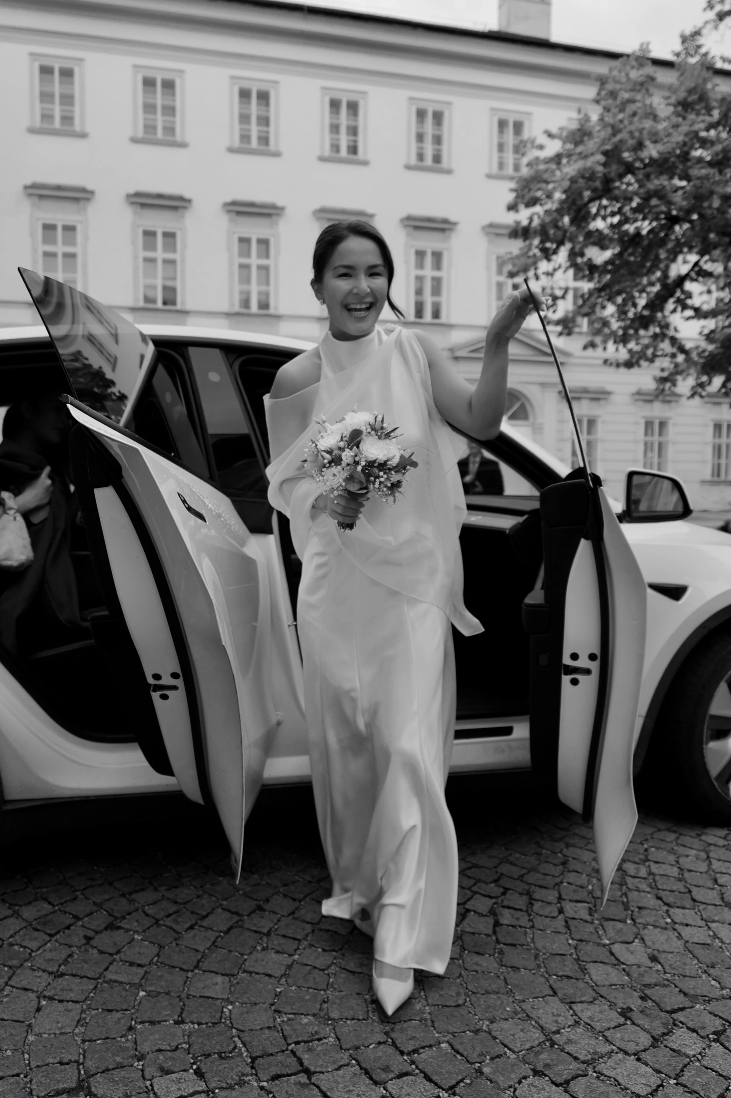 A woman in a wedding dress holding a bouquet of flowers, standing outside a vehicle with its door open, smiling and raising her arm. A tree and a building with multiple windows are visible in the background.