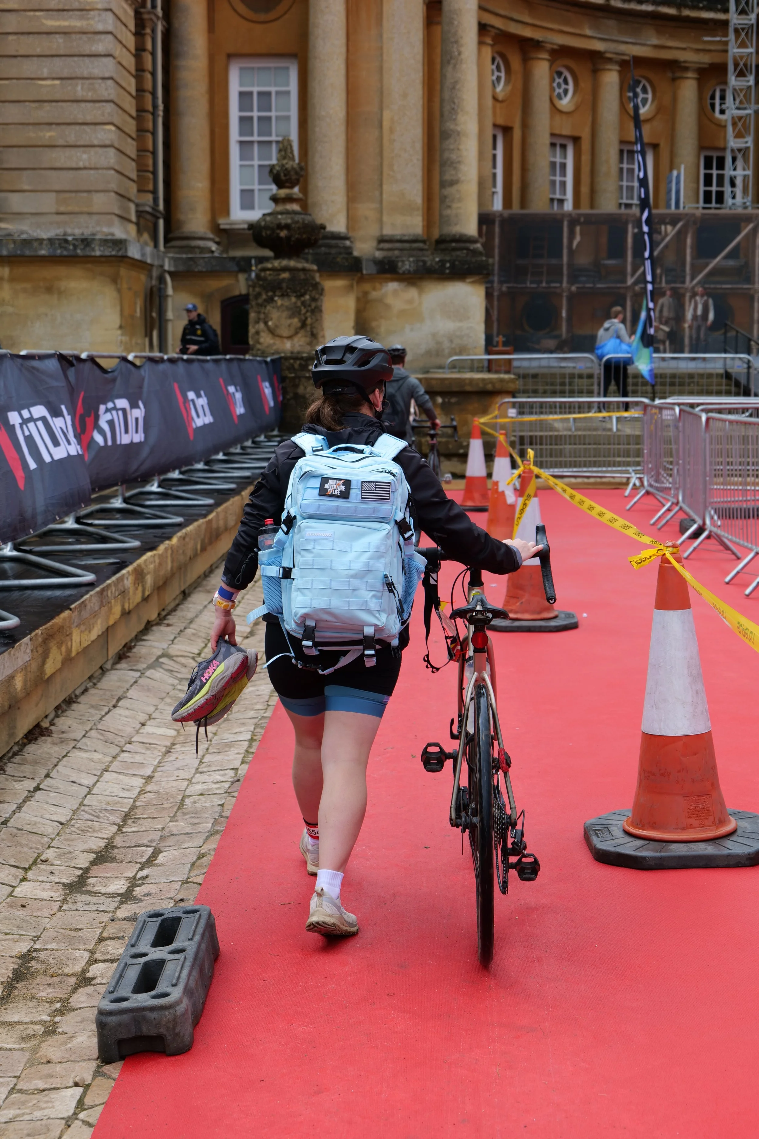 A woman walking with a bicycle on a red carpet outside a historic building, carrying a light blue backpack and holding a pair of shoes in her right hand, with cones and barriers around.