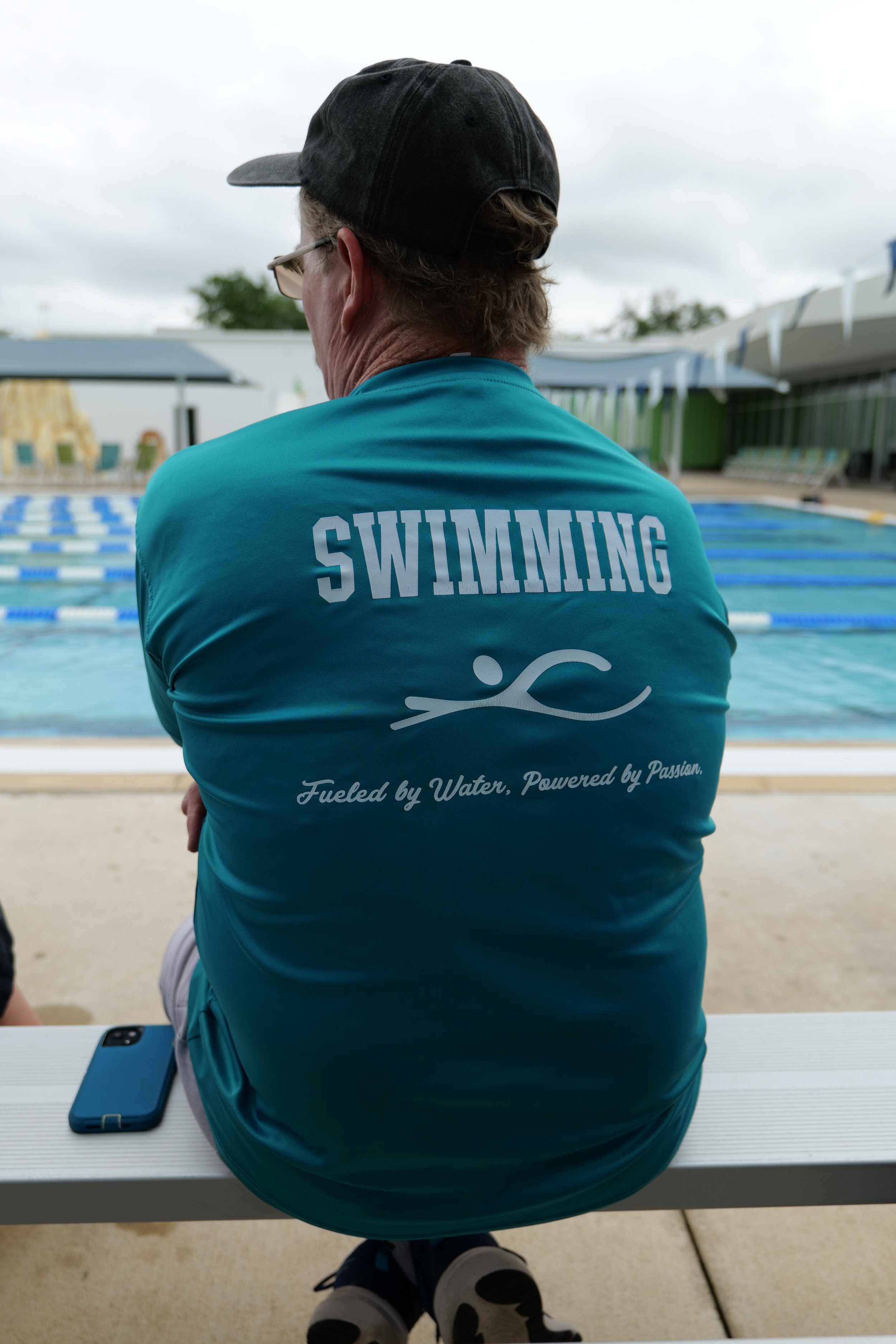 A person sitting at a swimming pool, wearing a blue T-shirt with 'SWIMMING' printed on the back and a swimmer icon, with a black cap and glasses, and a smartphone placed on the white table in front of them.