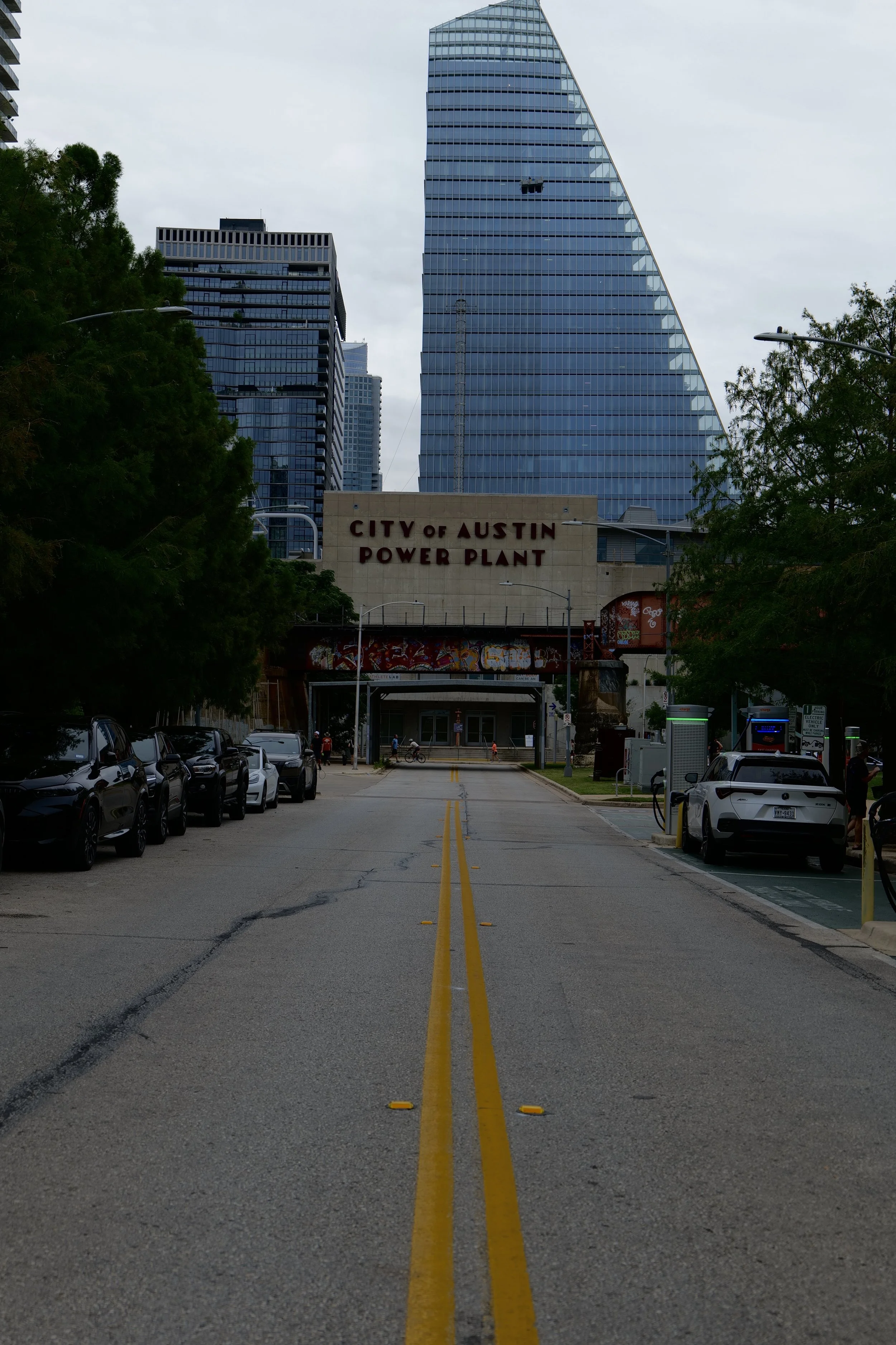 An urban street scene with a view of the City of Austin Power Plant sign in the foreground, tall modern glass skyscrapers in the background, parked cars along the sides, and a yellow double line in the middle of the street under an overcast sky.