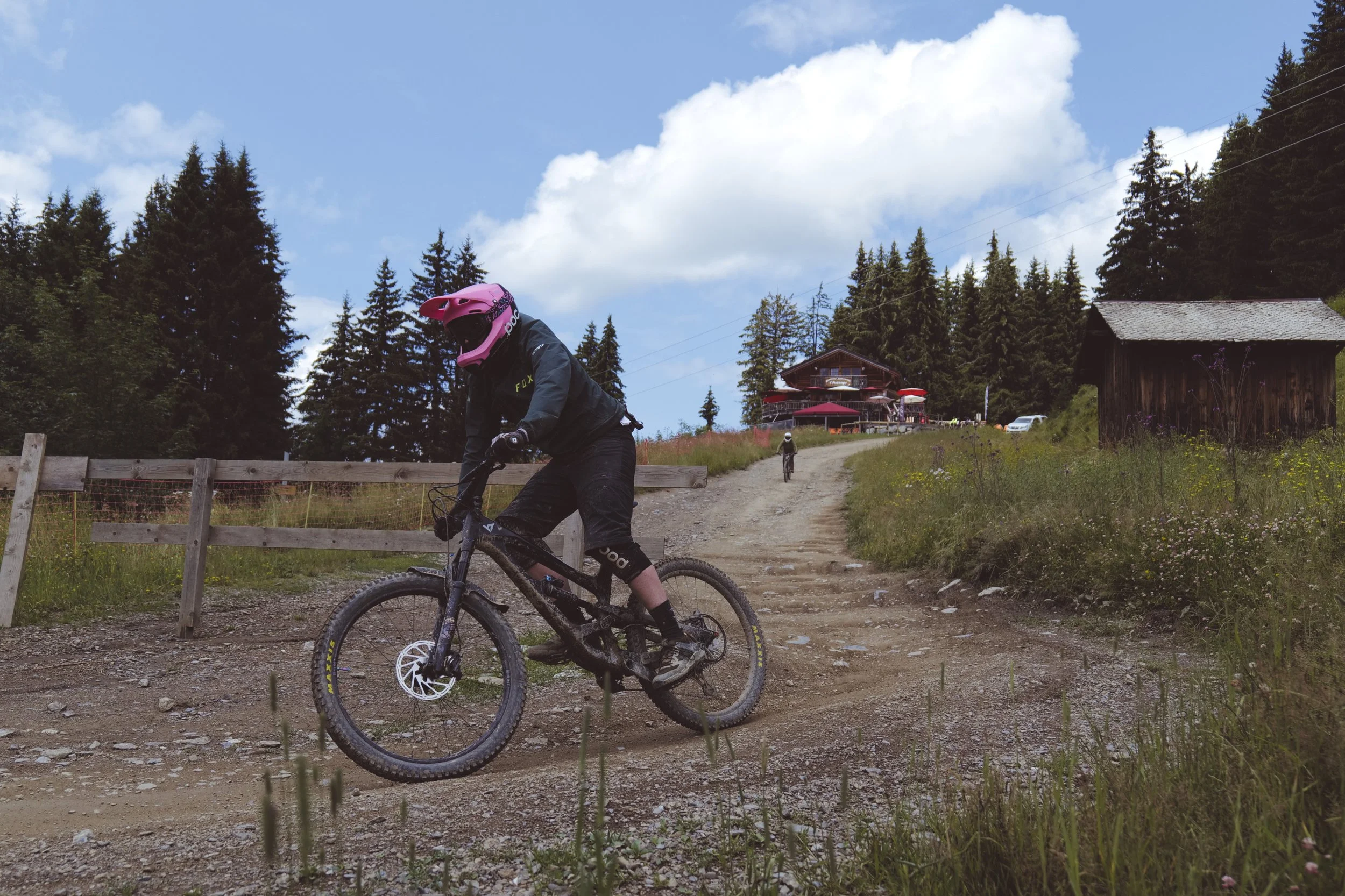 A mountain biker wearing a pink helmet, black jacket, and black shorts riding a bike on a dirt trail in a forested area with a house and trees in the background.
