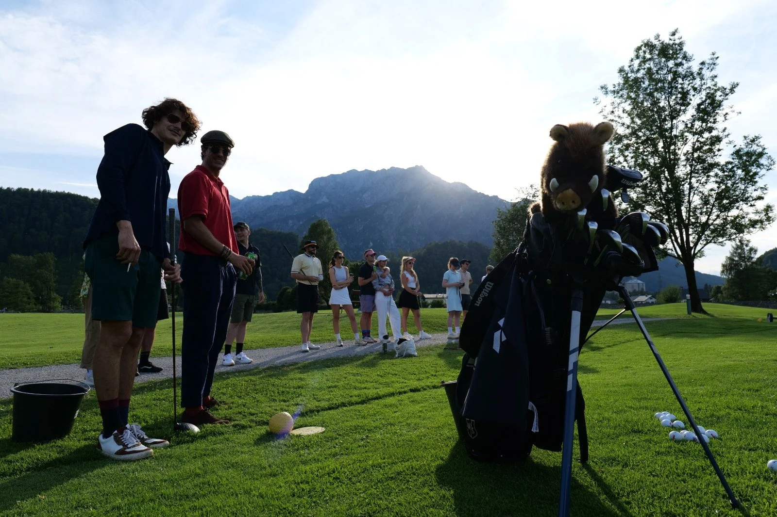 People gathered on a golf course with mountains in the background, some holding golf clubs, and a golf bag with a stuffed animal head in the foreground.
