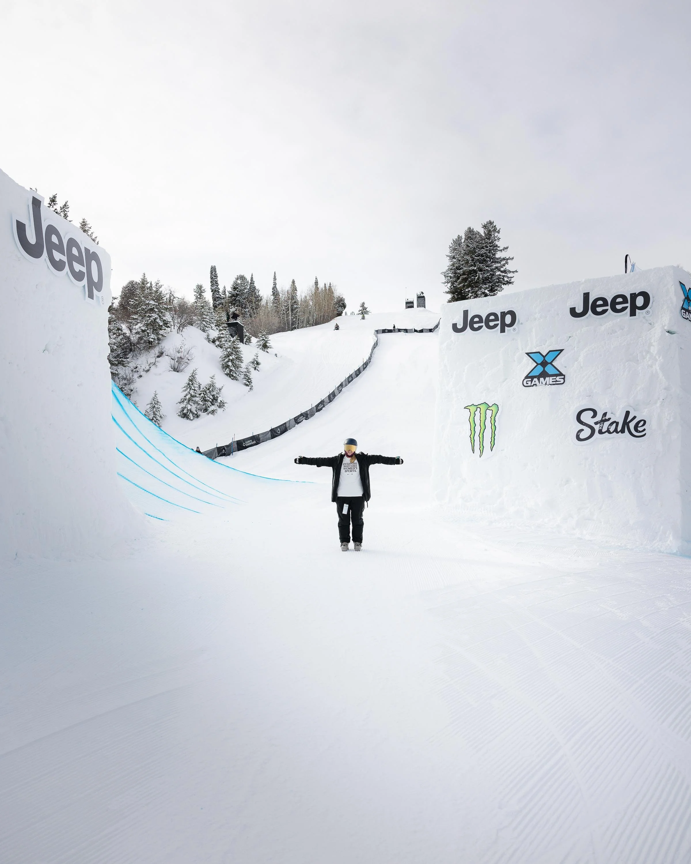 A person standing with arms outstretched on a snow-covered slope at a snowboarding or skiing competition, with prominent banners displaying logos such as Jeep, X Games, Monster, and Stake in the snow.