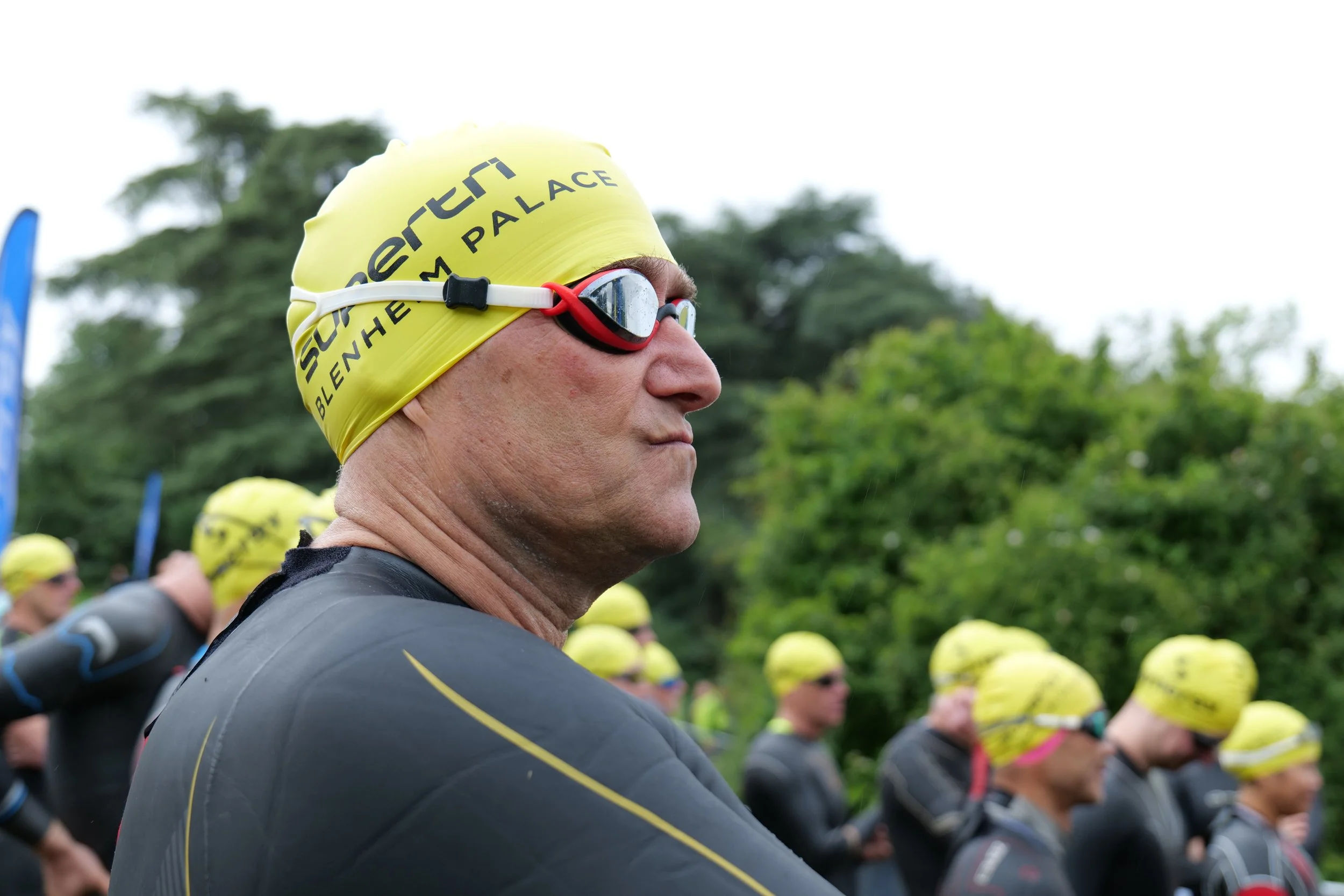 Close-up of a male athlete wearing a yellow swim cap and goggles, standing among a group of other swimmers with yellow caps outdoors.