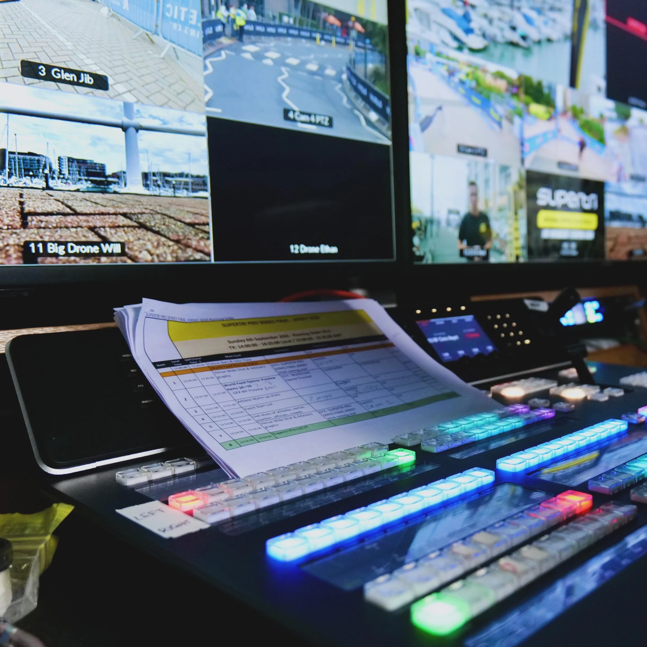 Multiple computer screens and a keyboard with colorful backlit keys, displaying various schedules, documents, and video feeds with a person visible on one monitor and a marina with boats outside the window.