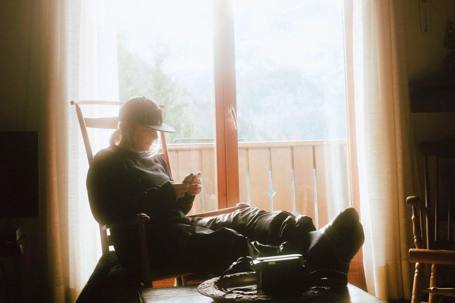 Person sitting on a wooden chair near a glass door, using a smartphone, with natural light coming through the window, and a wooden balcony visible outside.