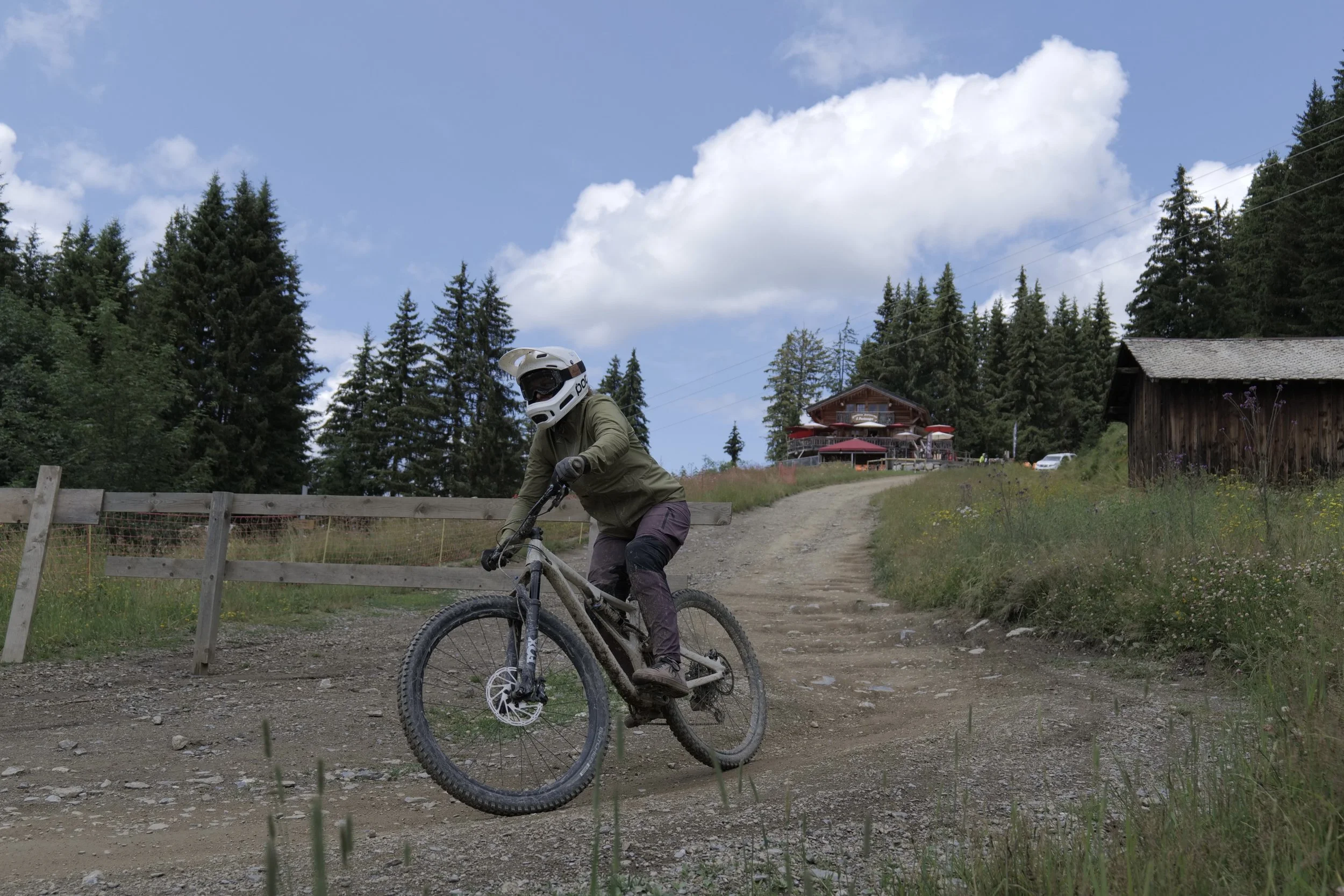 A person mountain biking on a dirt trail in a forested area with a building and trees in the background under a partly cloudy sky.