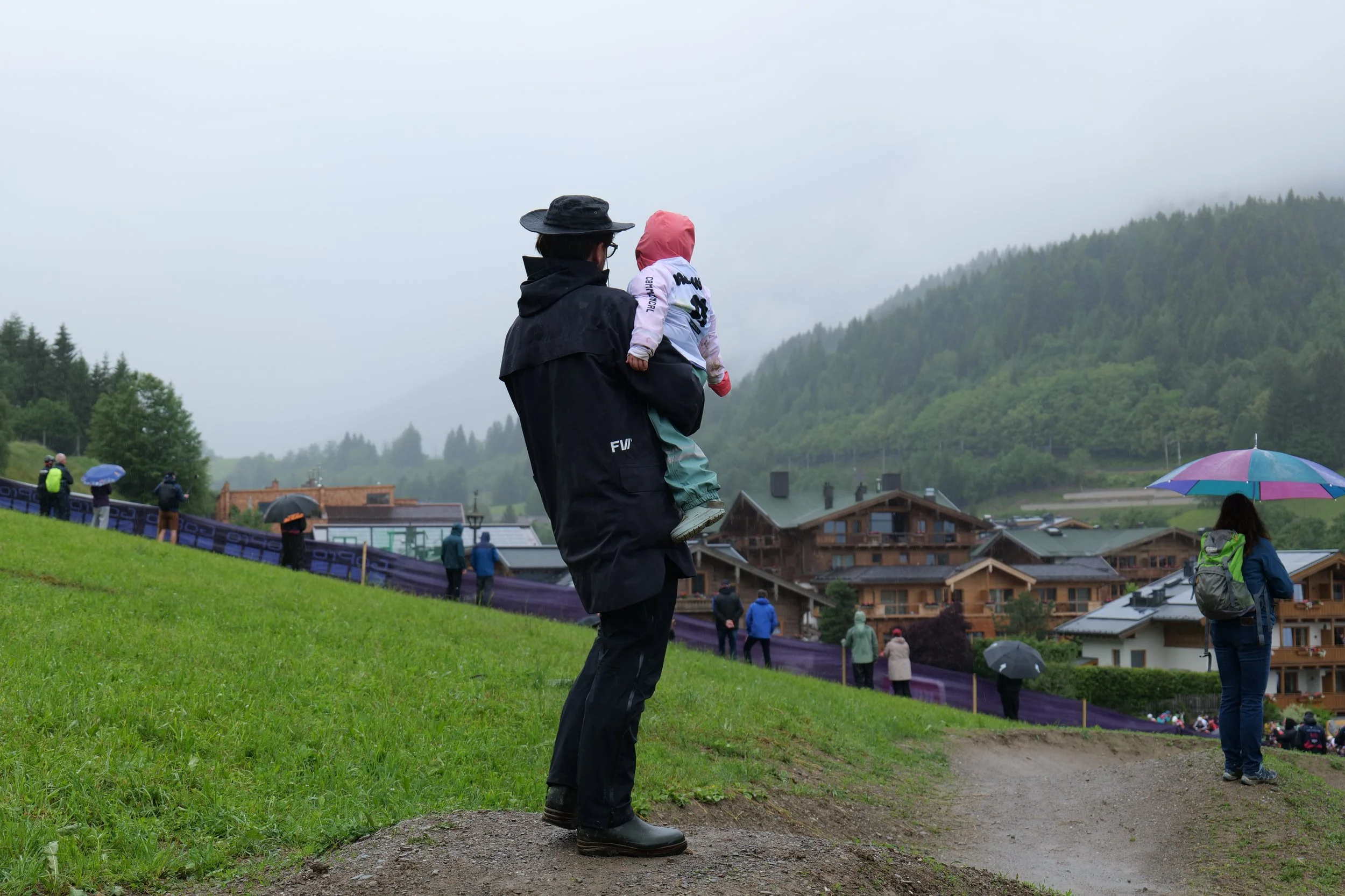 A person in rain gear holding a child with a pink hood at an outdoor event in a mountainous, foggy area, with other spectators and umbrellas visible.