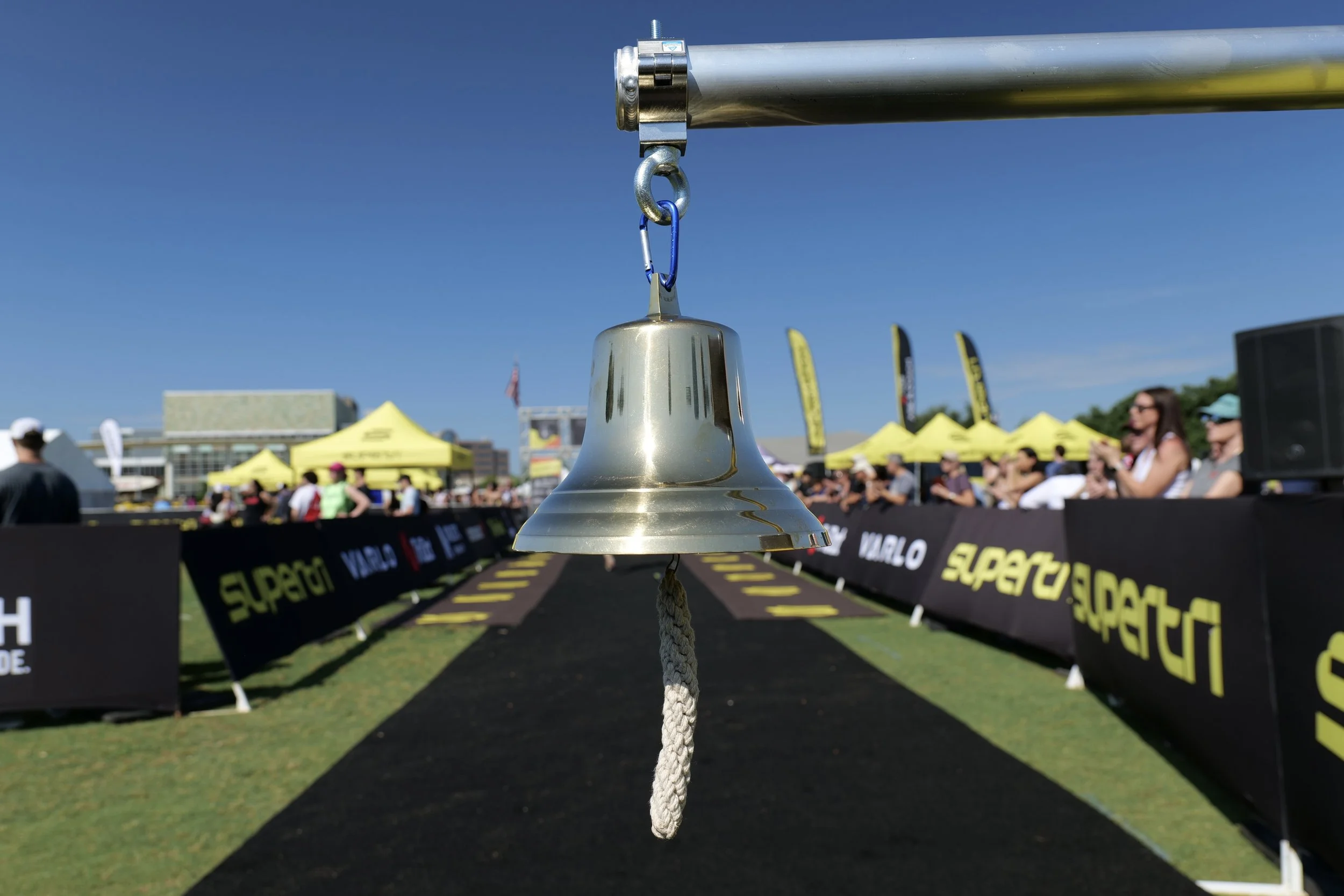 A metal bell hanging from a rope at an outdoor event with yellow tents and people in the background under a clear blue sky.