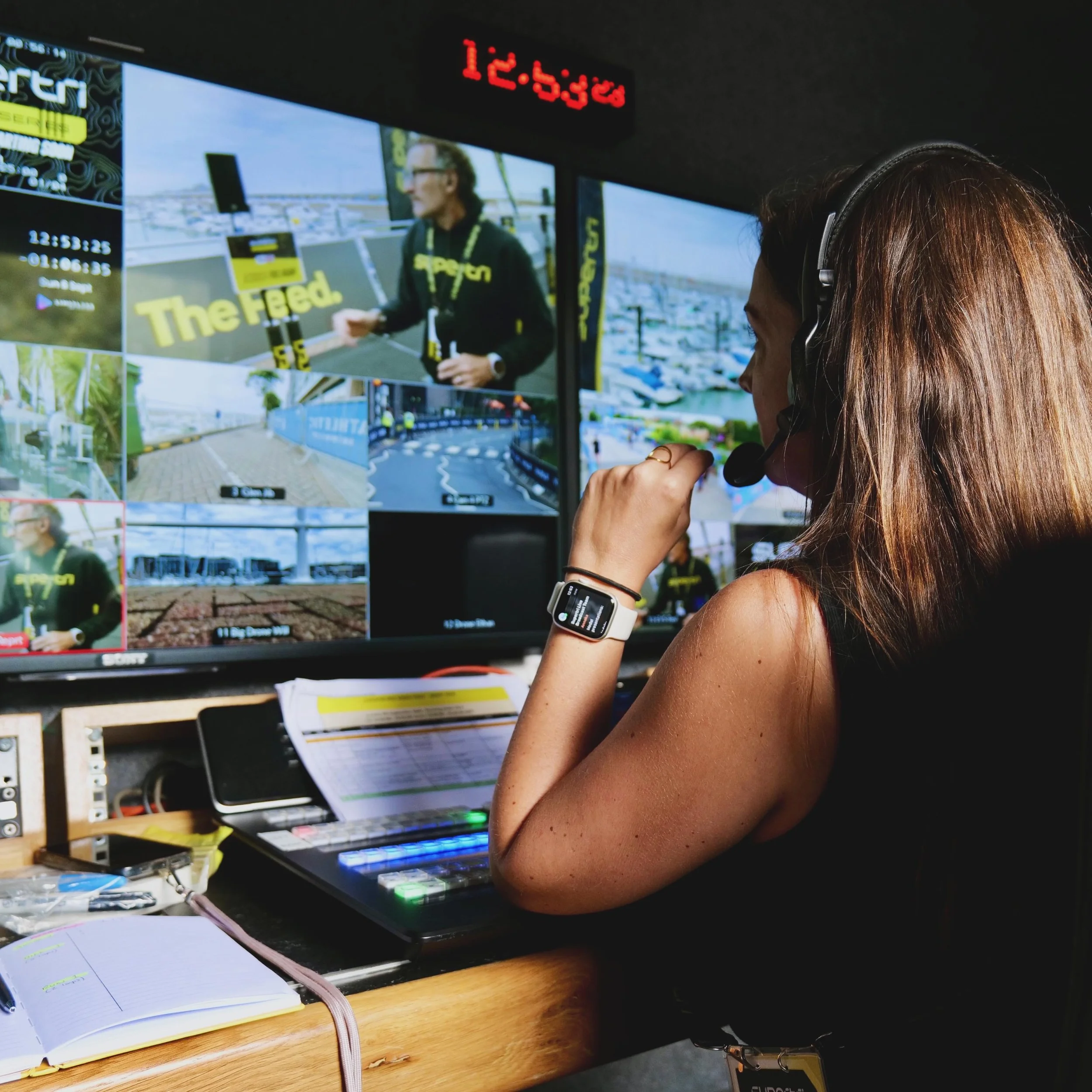 A person with long brown hair wearing headphones and a smartwatch, sitting at a desk with multiple monitors displaying a woman and various images, including a race track, at a broadcasting or editing station.