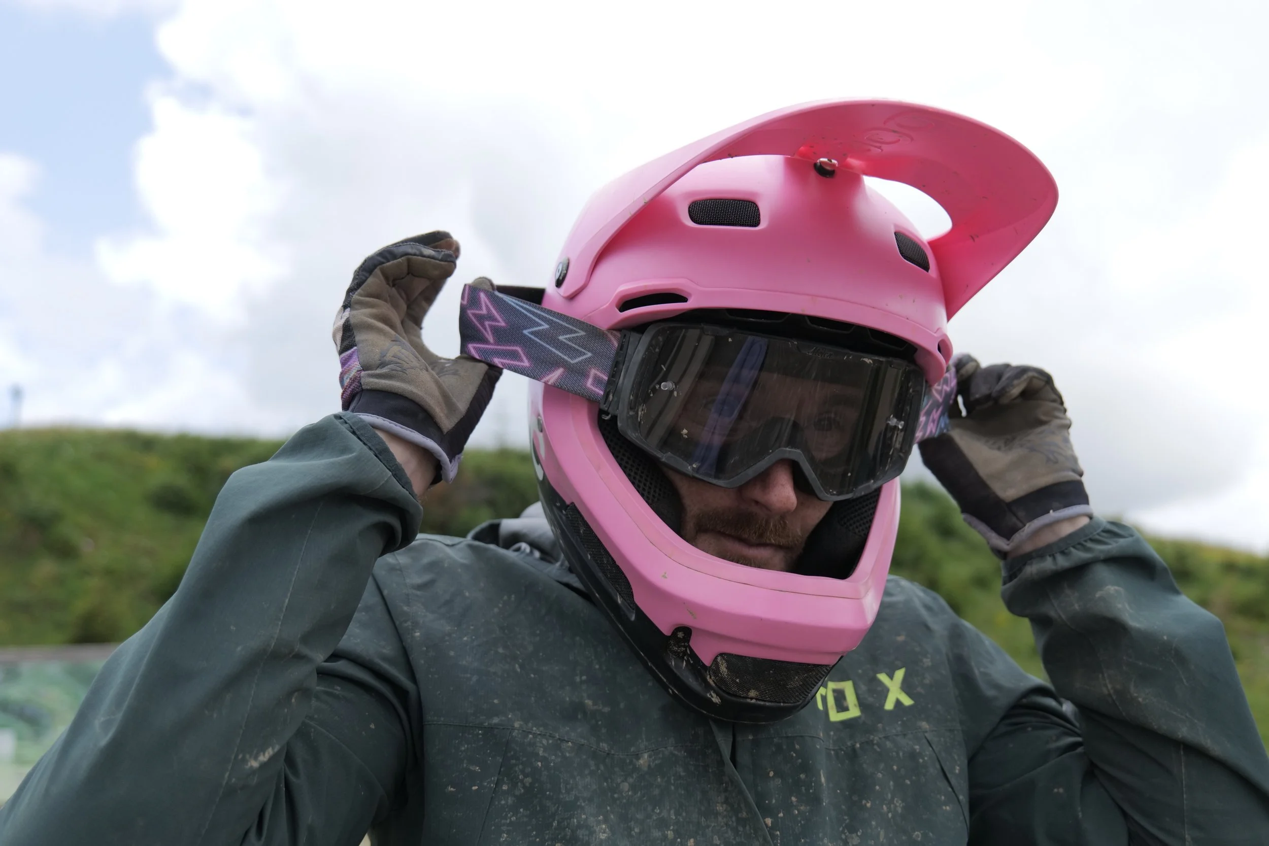A person in protective gear, including gloves and a pink helmet with a visor, is adjusting the helmet's strap outdoors on a cloudy day with green hills in the background.