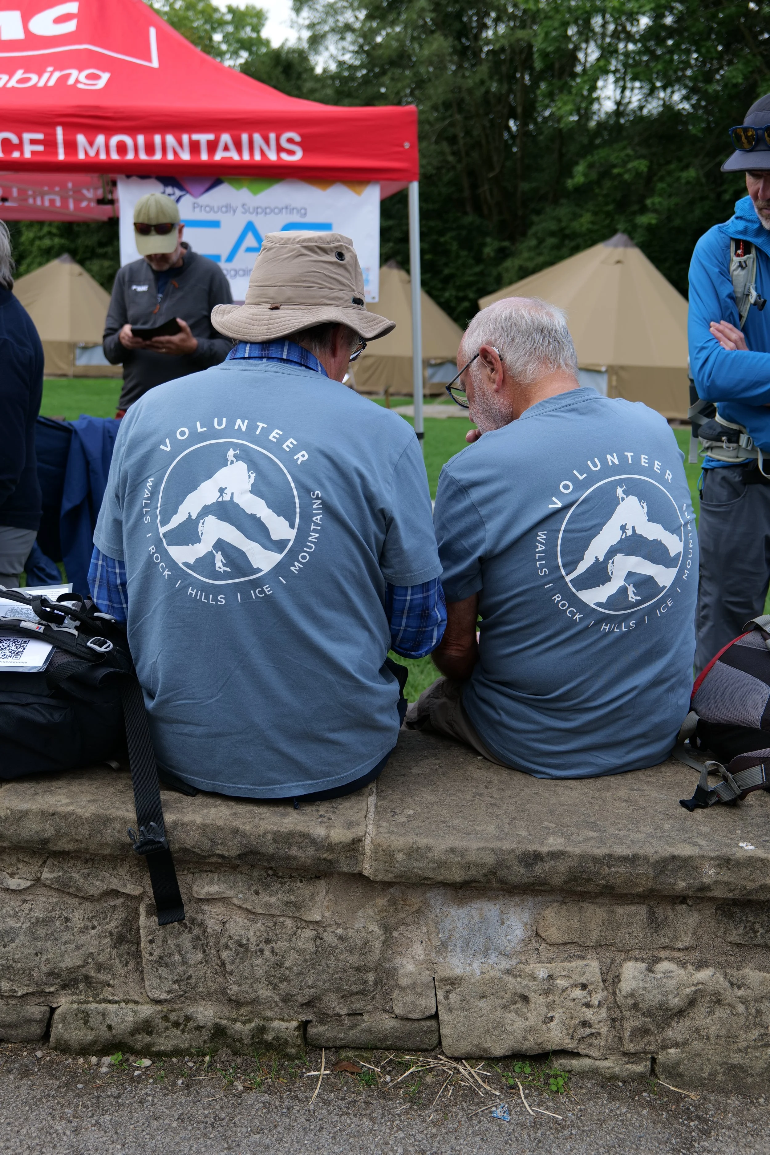Two people sitting on a stone ledge with their backs to the camera, wearing matching gray t-shirts with a mountain logo and the words 'Volunteer Wallis, Rock, Hills, Ice, Mountains.' They are outdoors, with tents and trees in the background. One pers