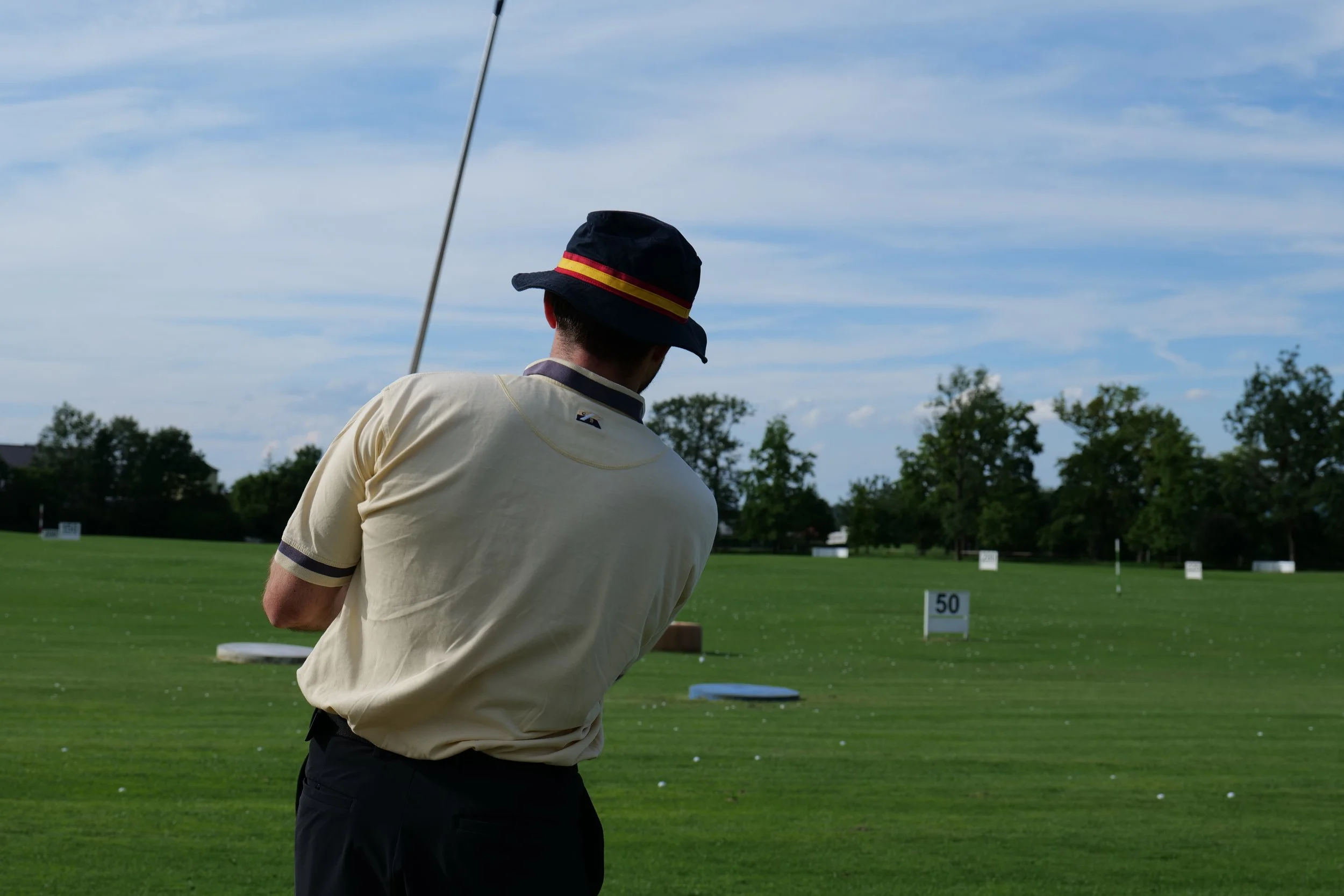 A man wearing a beige shirt and a black hat with a red and yellow band is practicing golf on a grassy field with white markers numbered 50 and 100, trees in the background, under a partly cloudy sky.