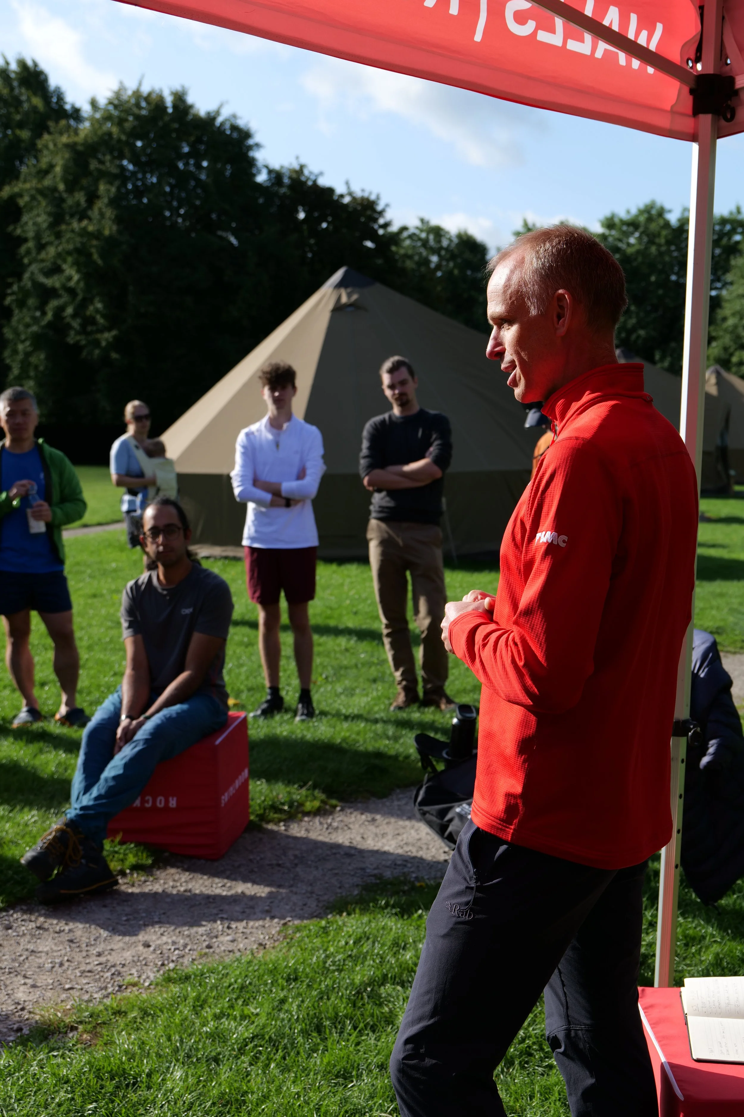 A man in a red jacket speaking outdoors at a gathering, with a group of people listening, tents and green trees in the background.