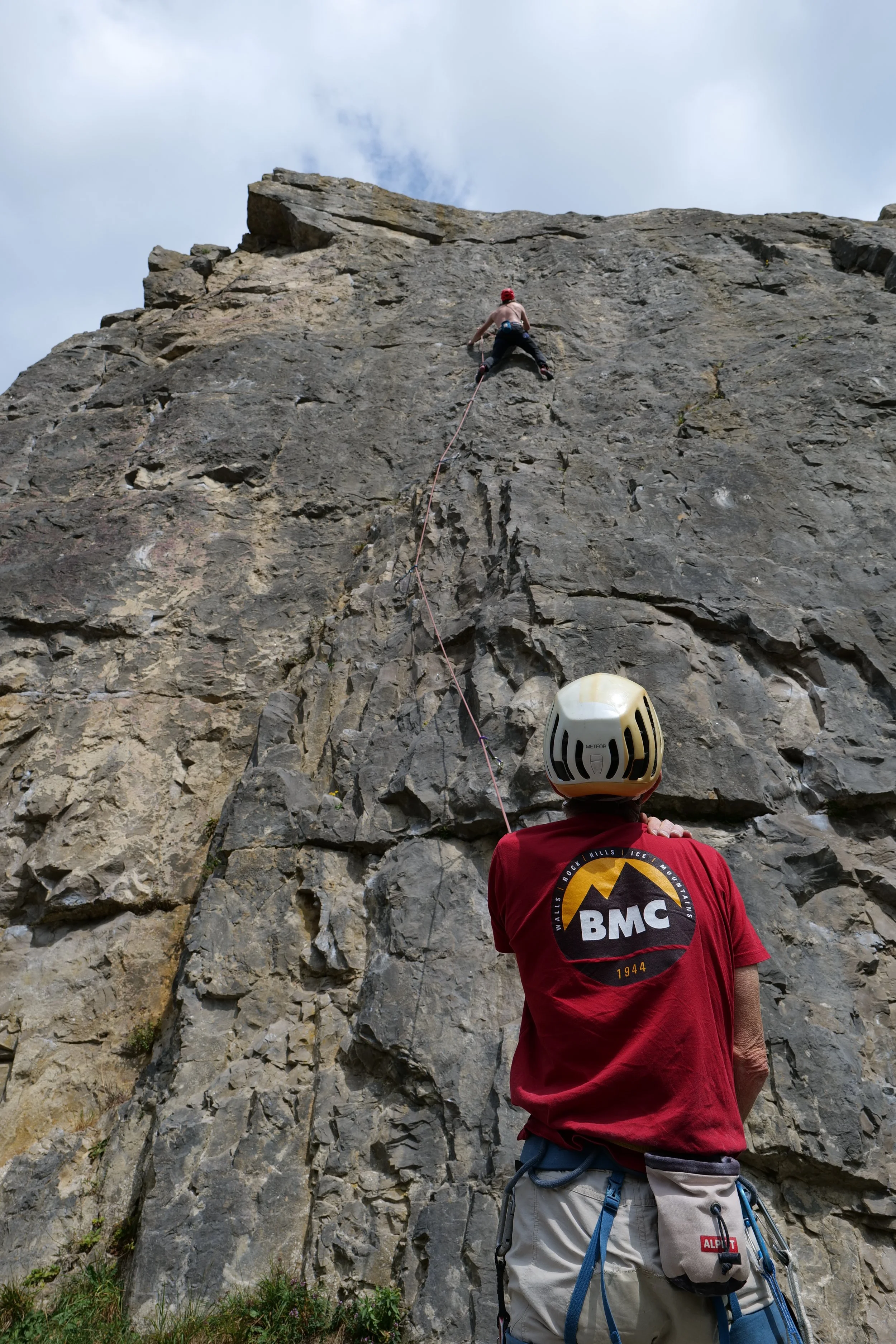 A rock climber ascending a steep rock face with another person observing from below during daytime.