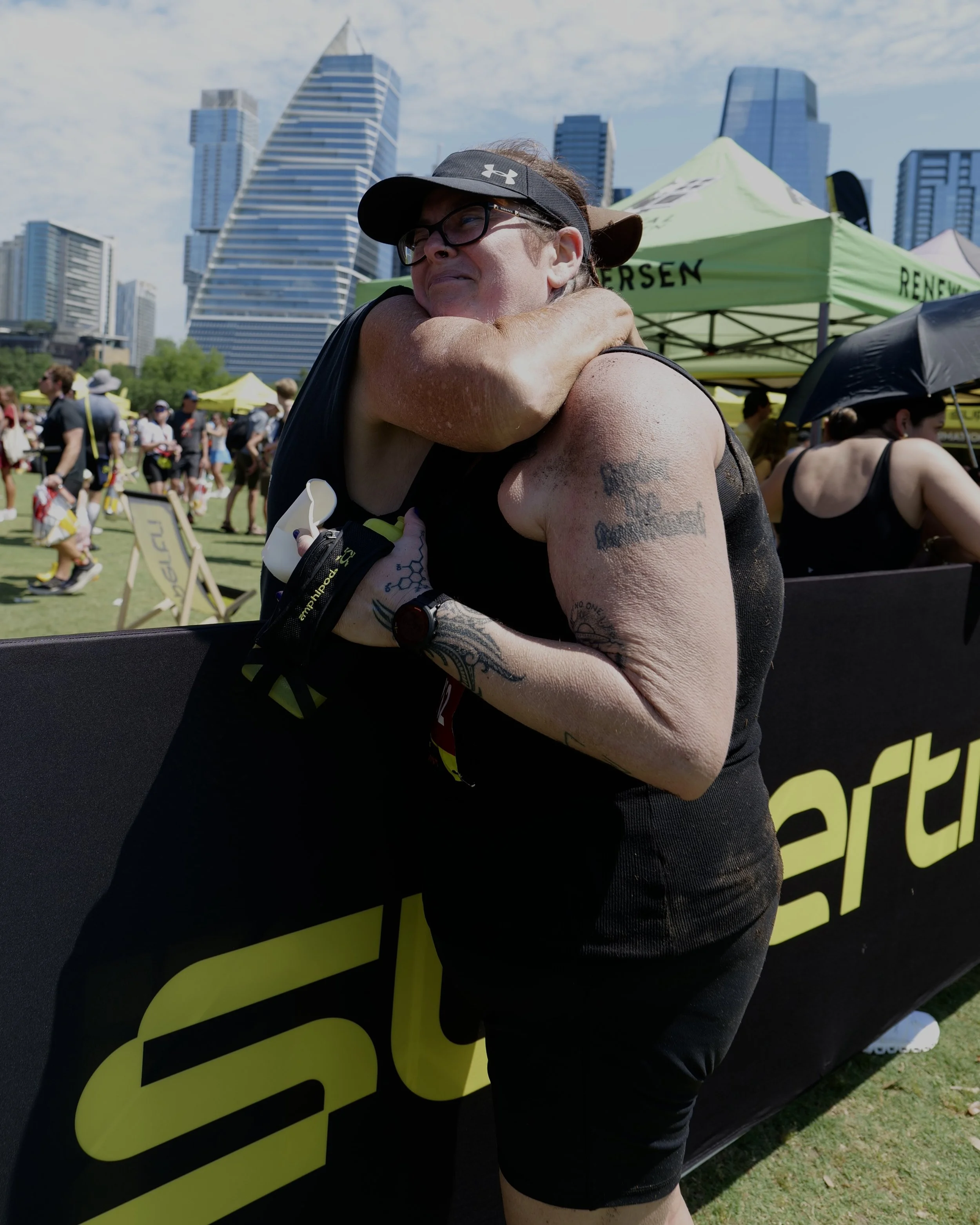 A woman with glasses and a black visor hugs another person at an outdoor event, with tents and tall buildings in the background.