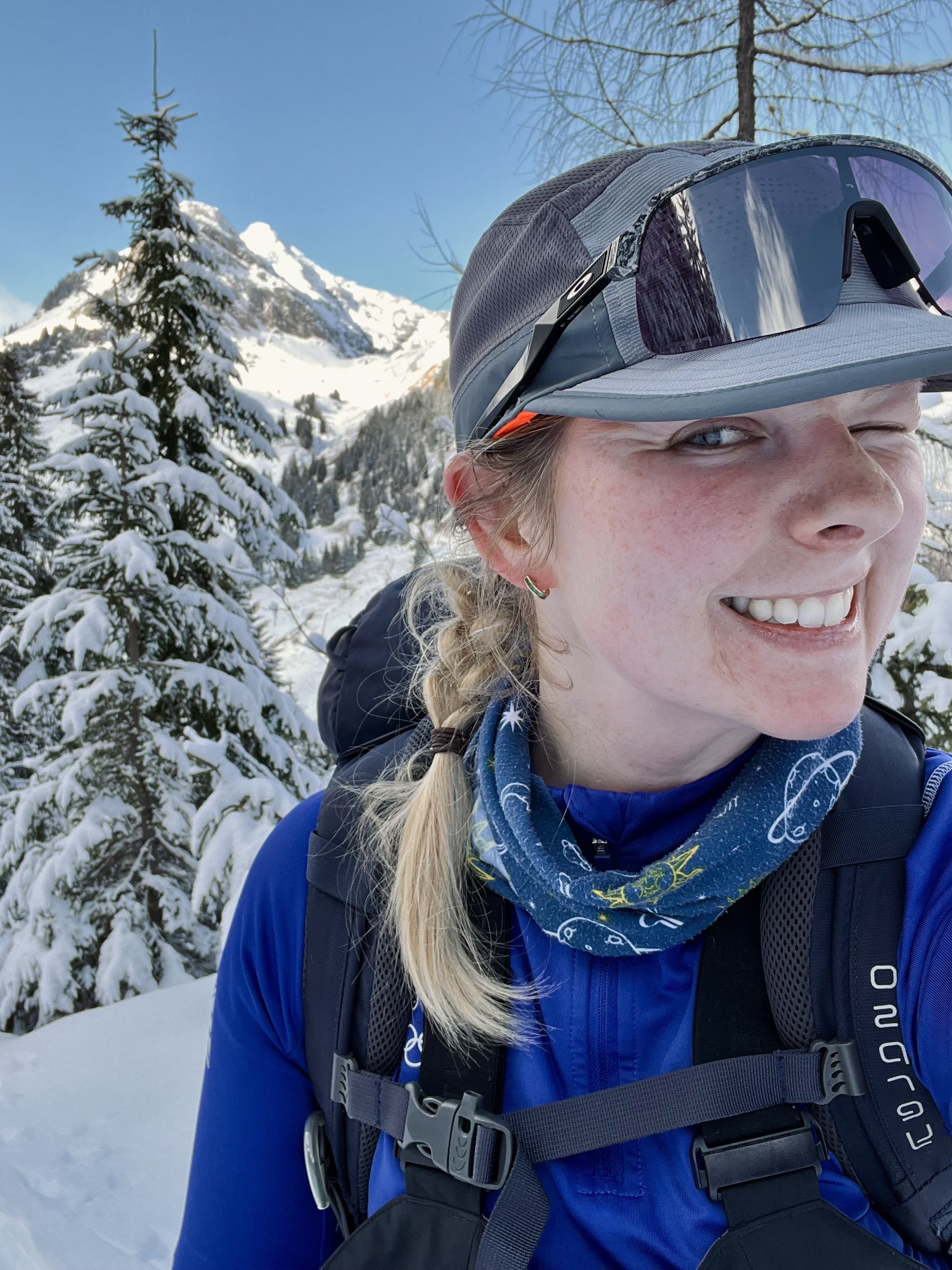 A woman in winter gear smiling in a snowy mountain landscape, wearing a grey cap with sunglasses on top, a blue jacket, and a backpack.