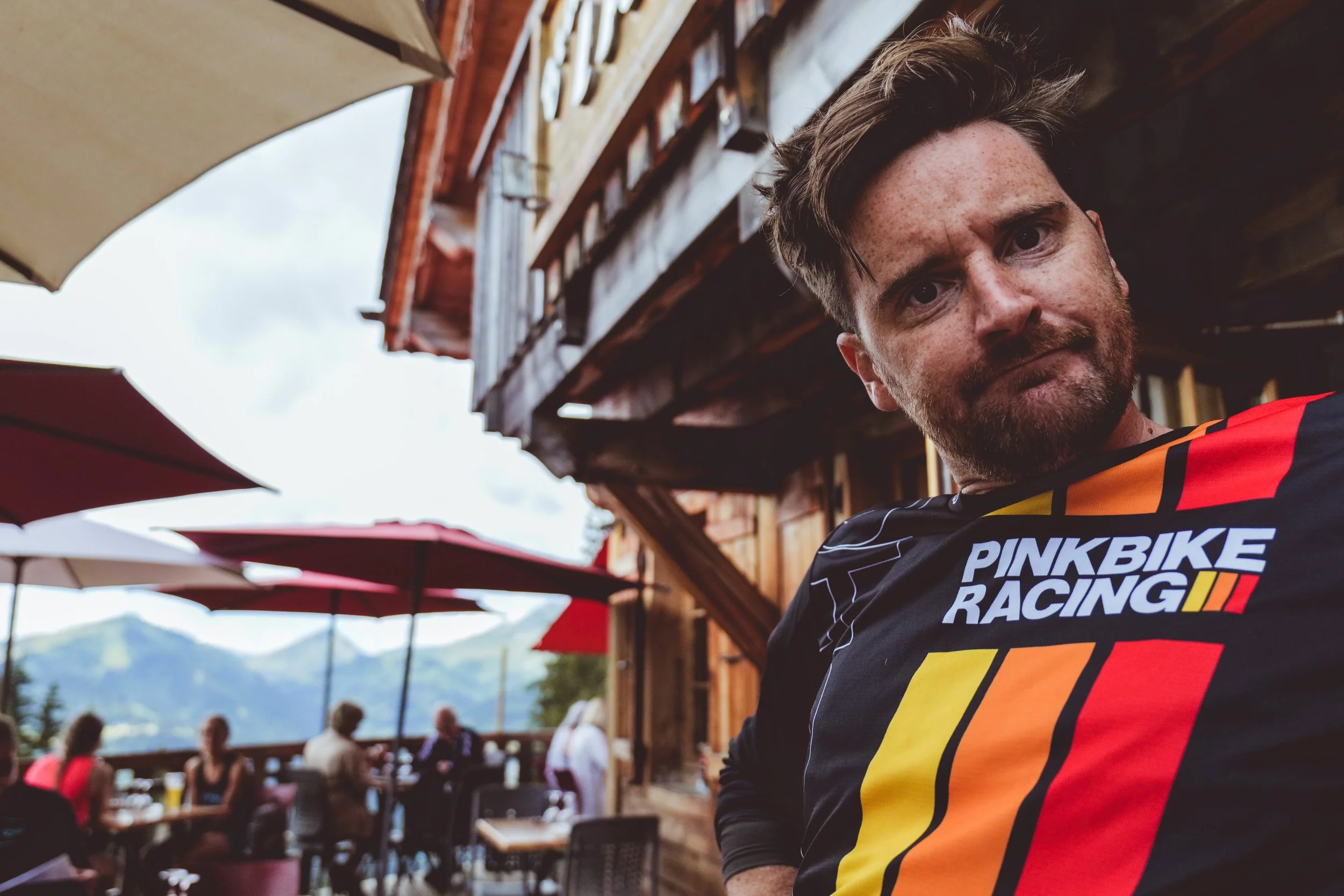 A man with brown hair and a beard wearing a Pinkbike Racing shirt, looking over his shoulder at the camera, with an outdoor restaurant or cafe setting with umbrellas, tables, and people in the background.