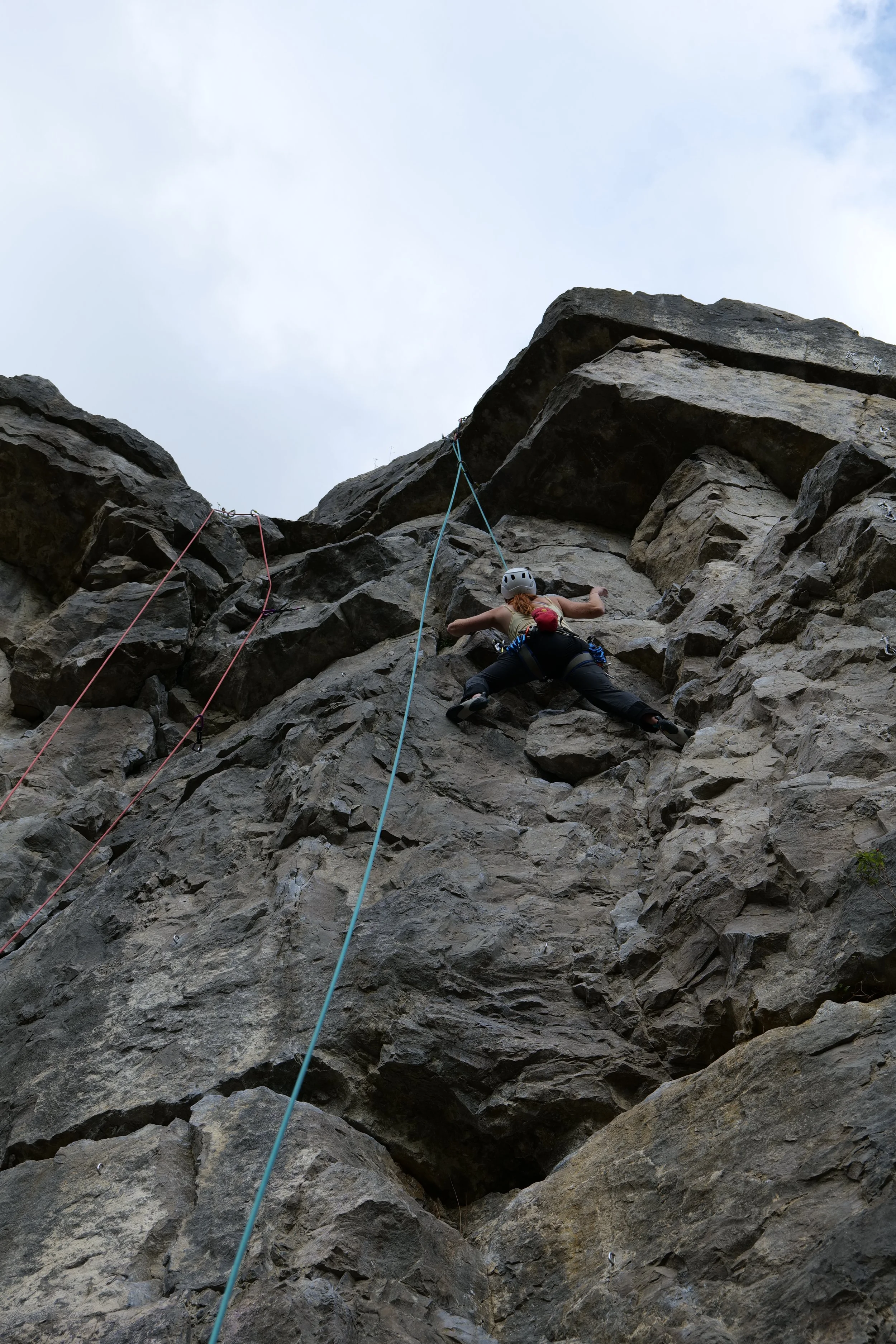 Person rock climbing on a steep, rocky cliff face with safety harness and helmet, viewed from below against a cloudy sky.