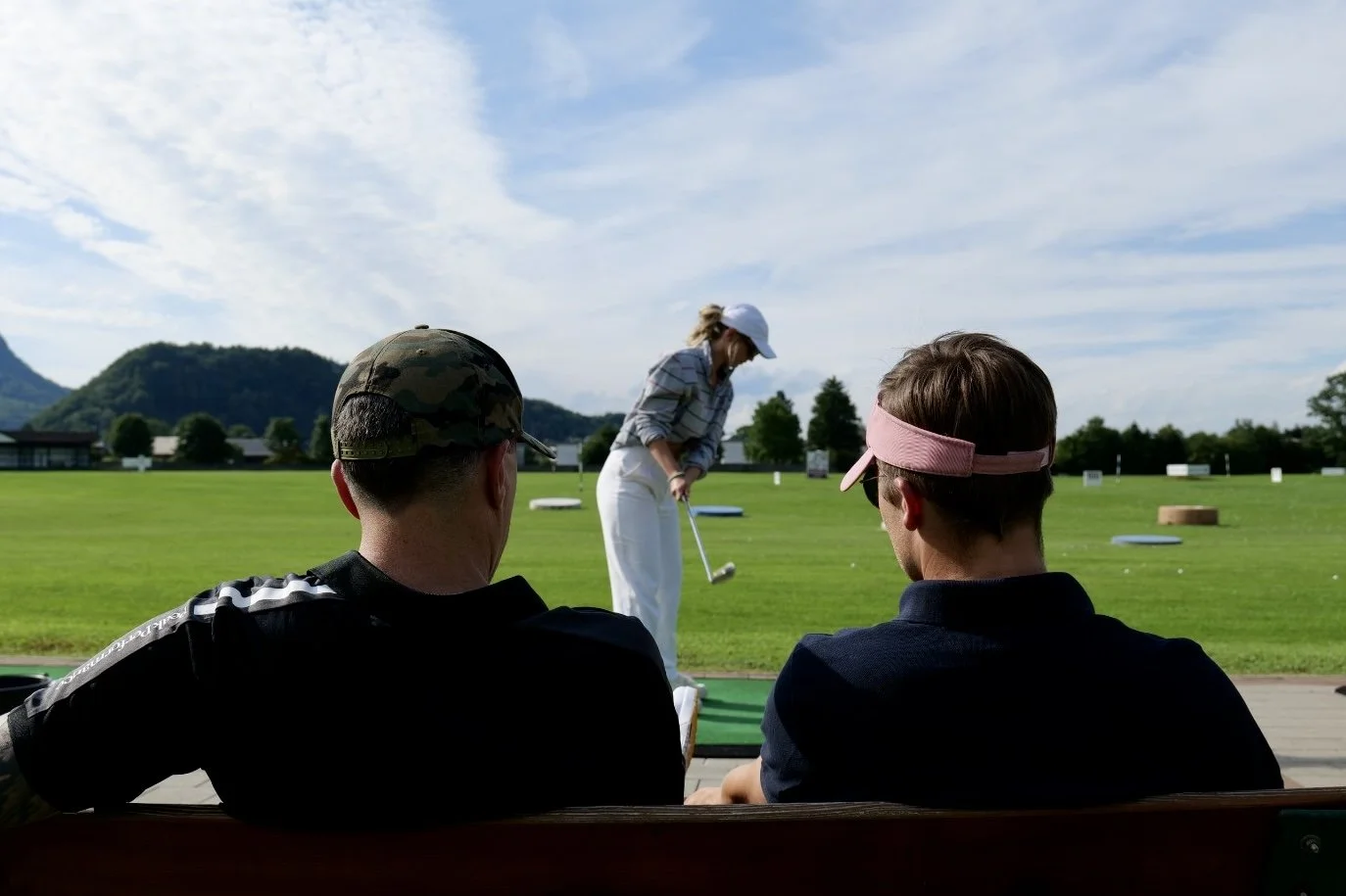 Two men sitting on a bench watching a woman practice golf on a driving range, with mountains and a blue sky in the background.