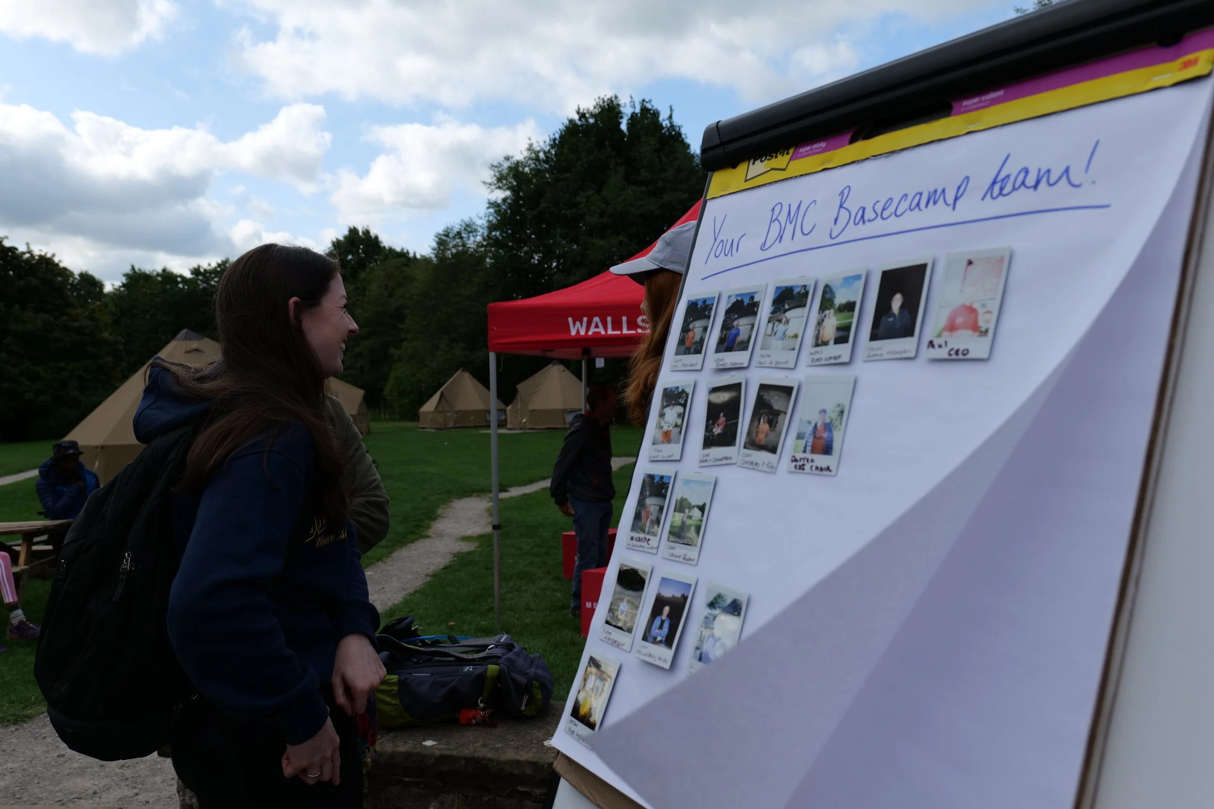 A woman is standing outdoors looking at a display board with photos and notes. There are tents and trees in the background, and a red tent with the word 'WALLS' is visible. The sky is partly cloudy.