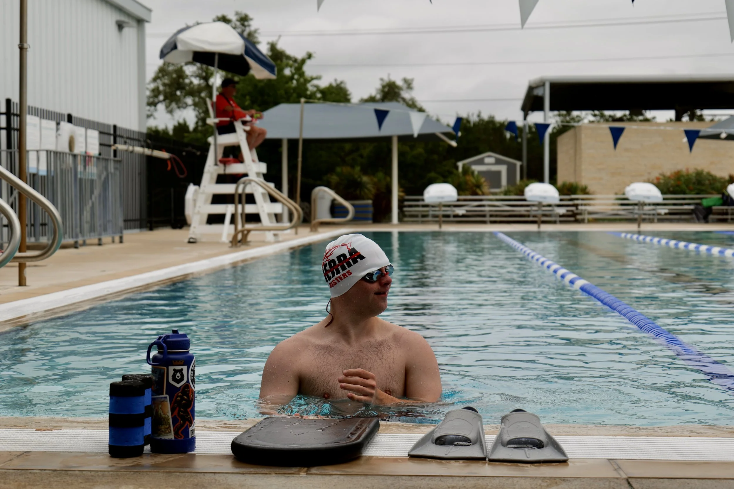 A swimmer wearing a white swim cap and goggles sitting in a swimming pool, with green water bottles and personal items on a nearby bench.
