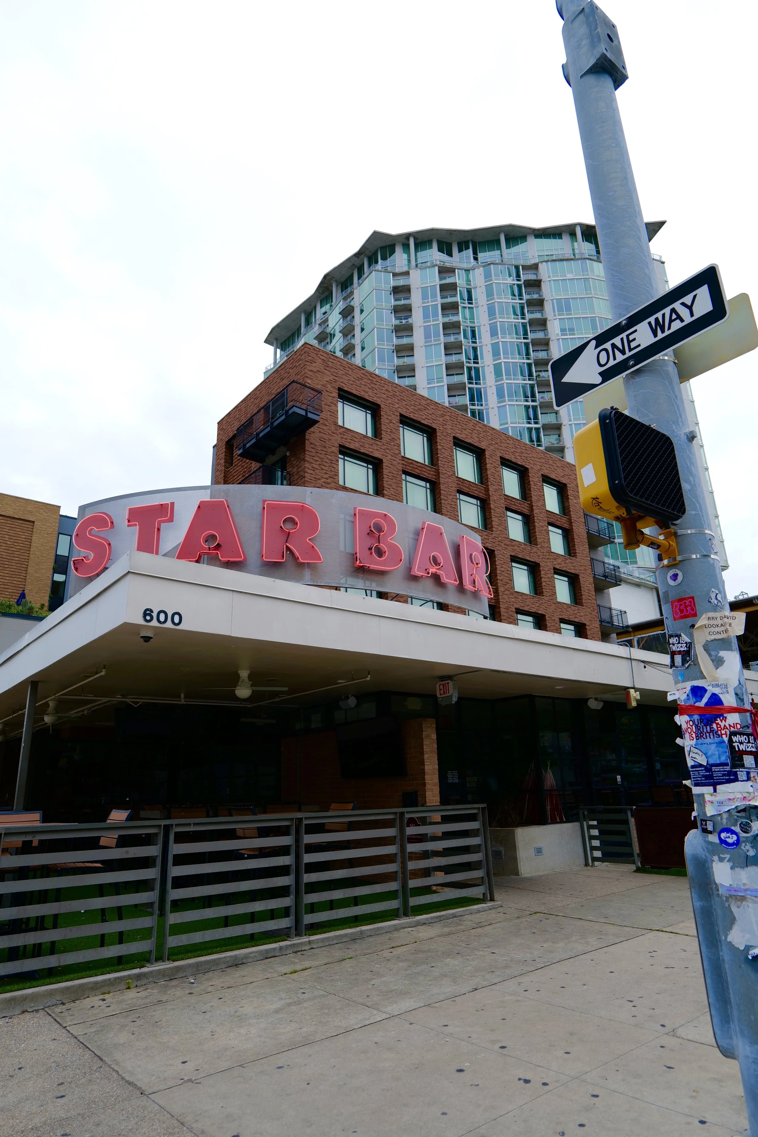 Street view showing a building with a red brick section and a taller glass skyscraper in the background. There is a neon sign reading 'STAR BAR' and a one-way street sign attached to a street pole. The sidewalk is visible with a security camera and a