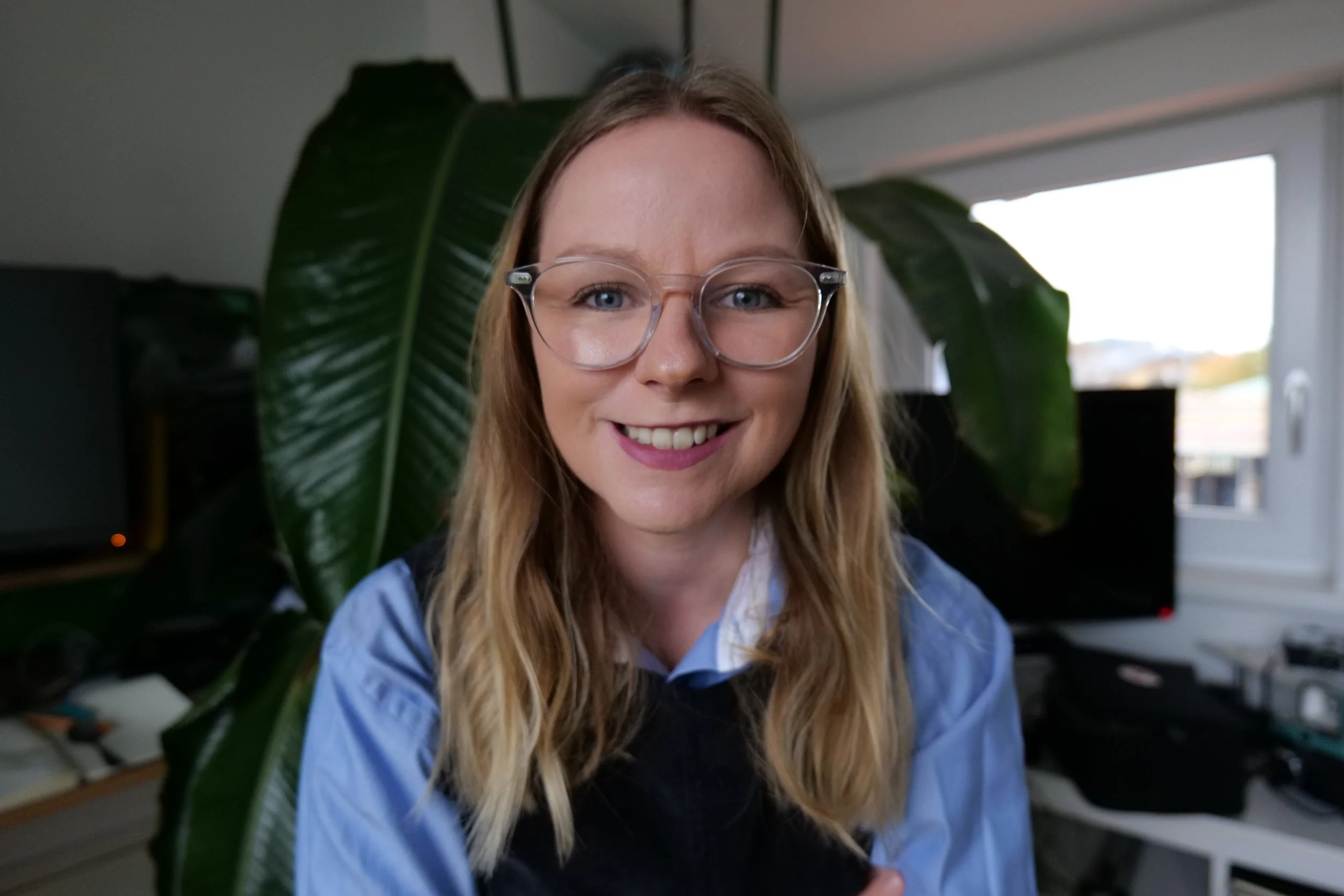 A woman with long blonde hair and glasses smiling in a room with a large green plant behind her and a window in the background.