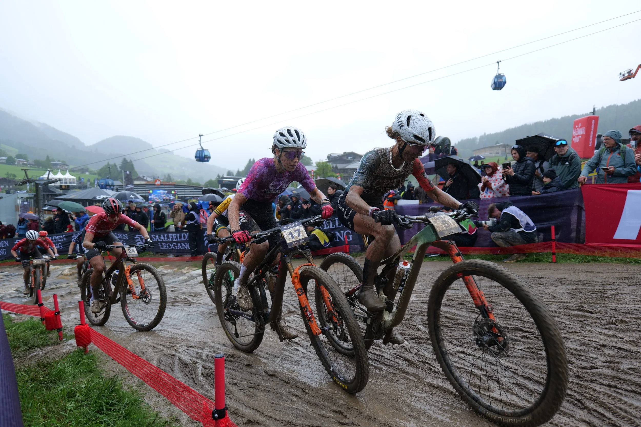 Cyclists in a mountain biking race on a muddy trail with spectators and gondolas in the background on a rainy day.