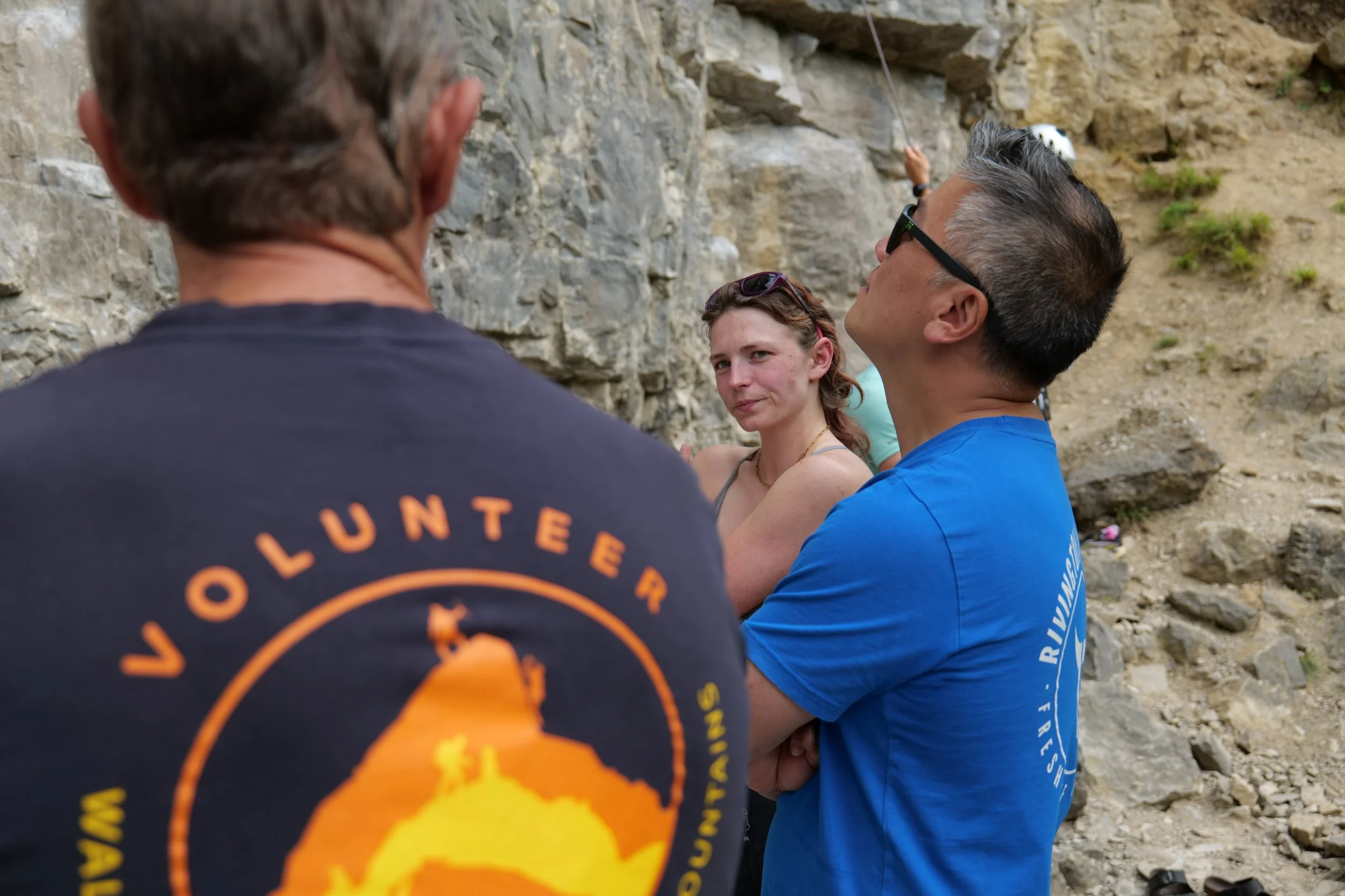 Three people are sitting on a rocky outdoor area during the daytime. One person is facing away from the camera, wearing a dark shirt with orange text. The other two are facing toward the camera; one is a young woman with reddish hair and sunglasses o