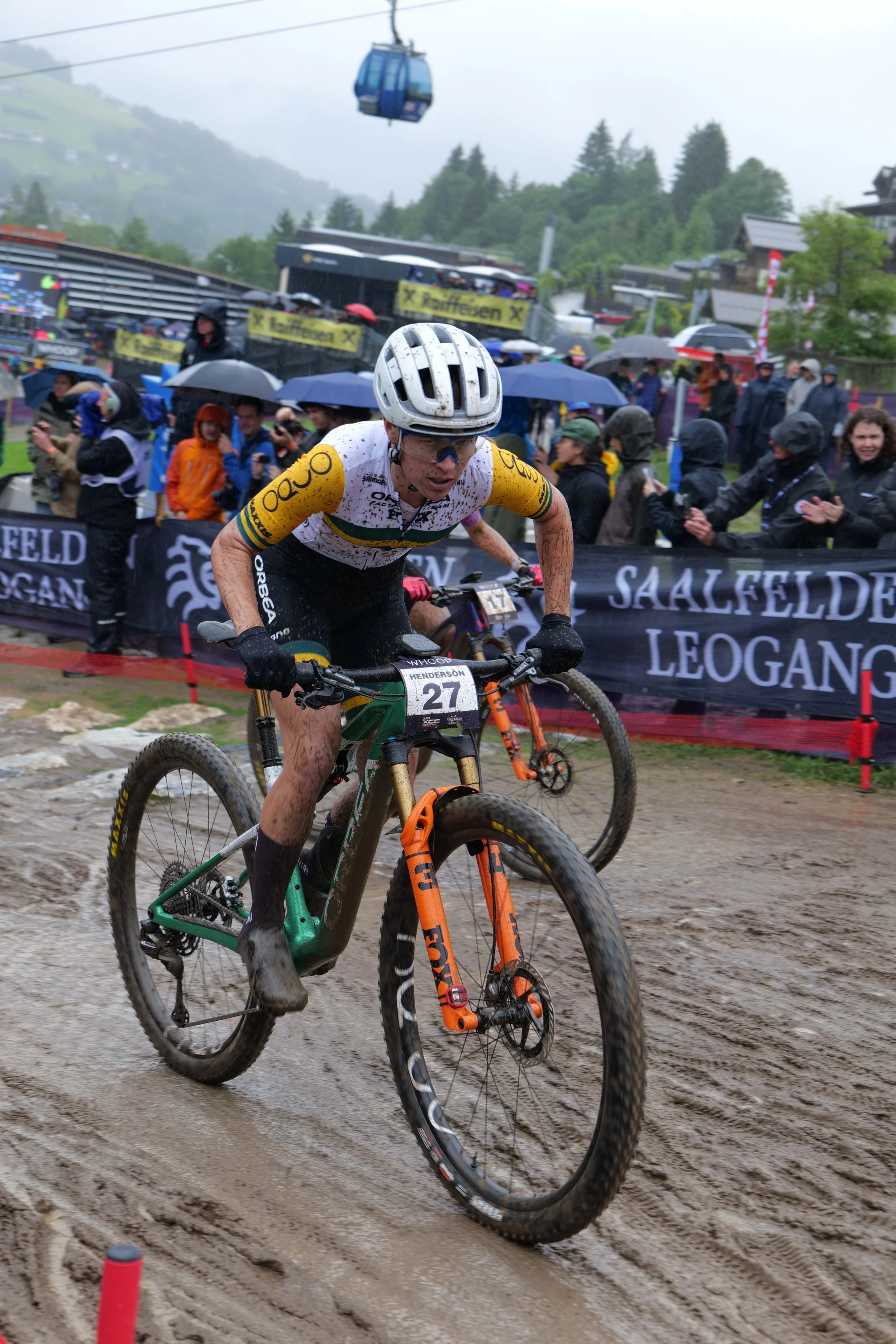 A cyclist in a helmet and dirt bike gear riding through a muddy course during a mountain biking race, with spectators and rain in the background.
