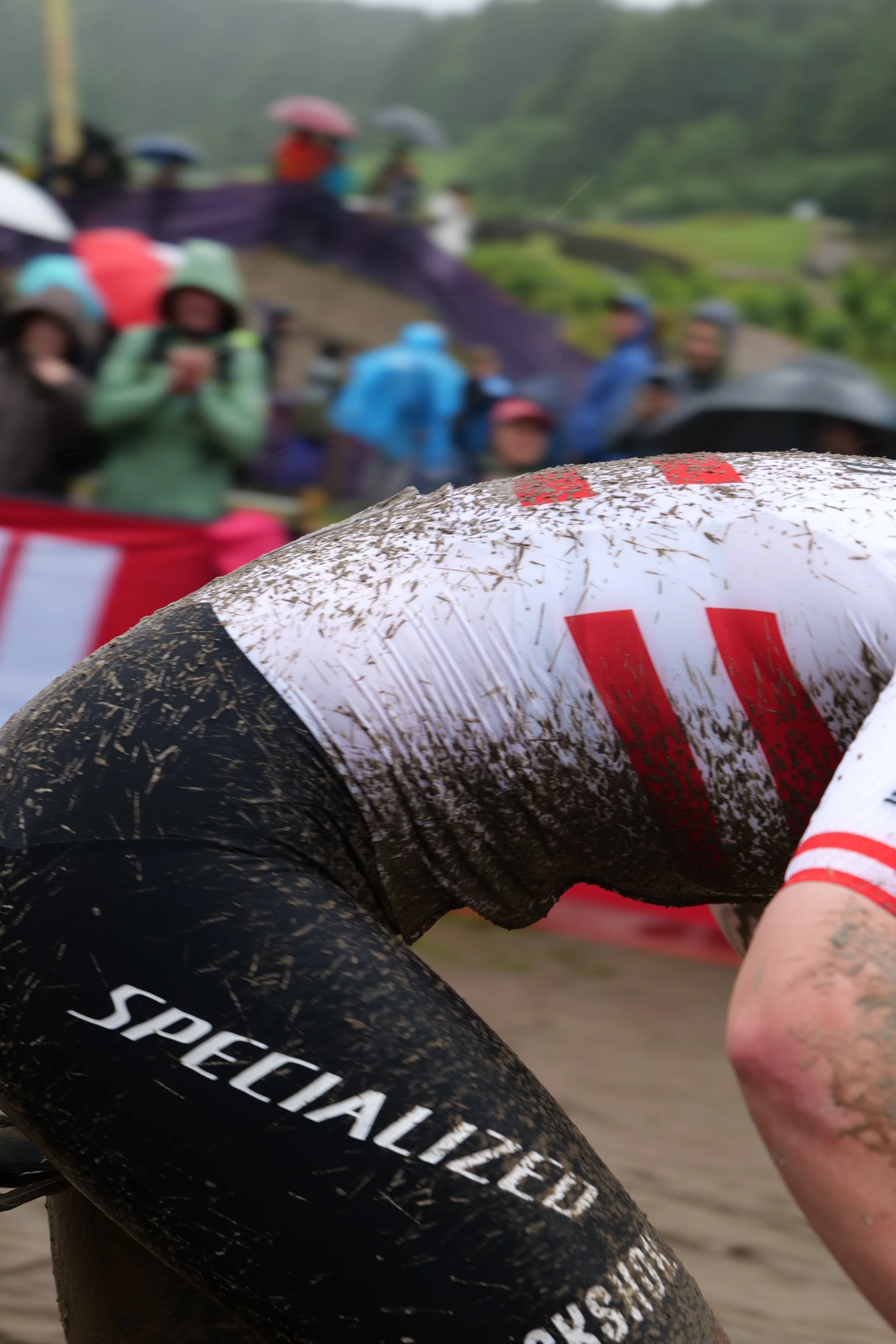 Close-up of a cyclist's muddy leg and arm during a race, with a blurred background of spectators with umbrellas on a rainy day.