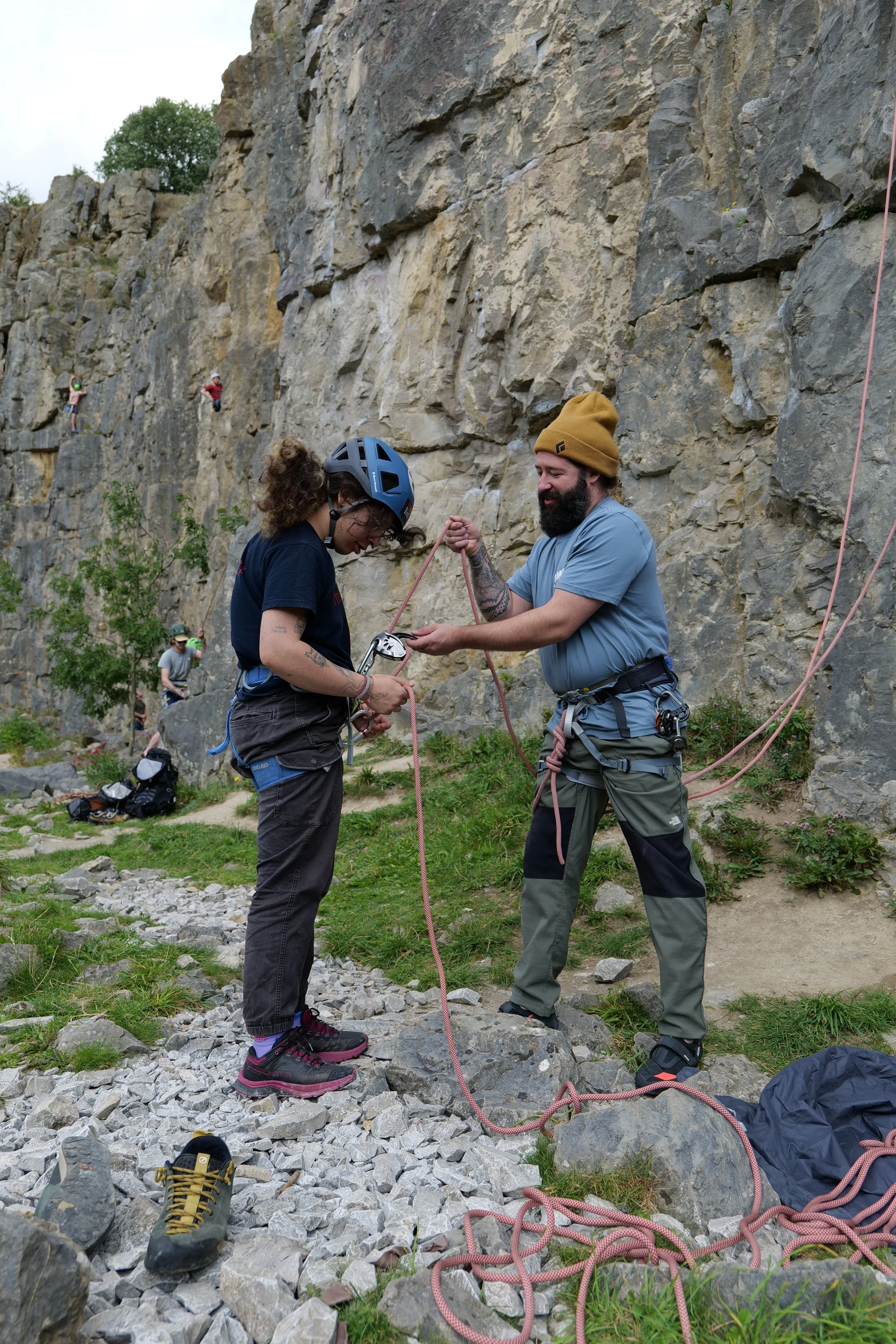 A woman wearing a helmet is preparing to belay a male climber on a rocky outdoor cliff with multiple climbers in the background. The man is holding the climbing rope, and climbing shoes are on the ground.