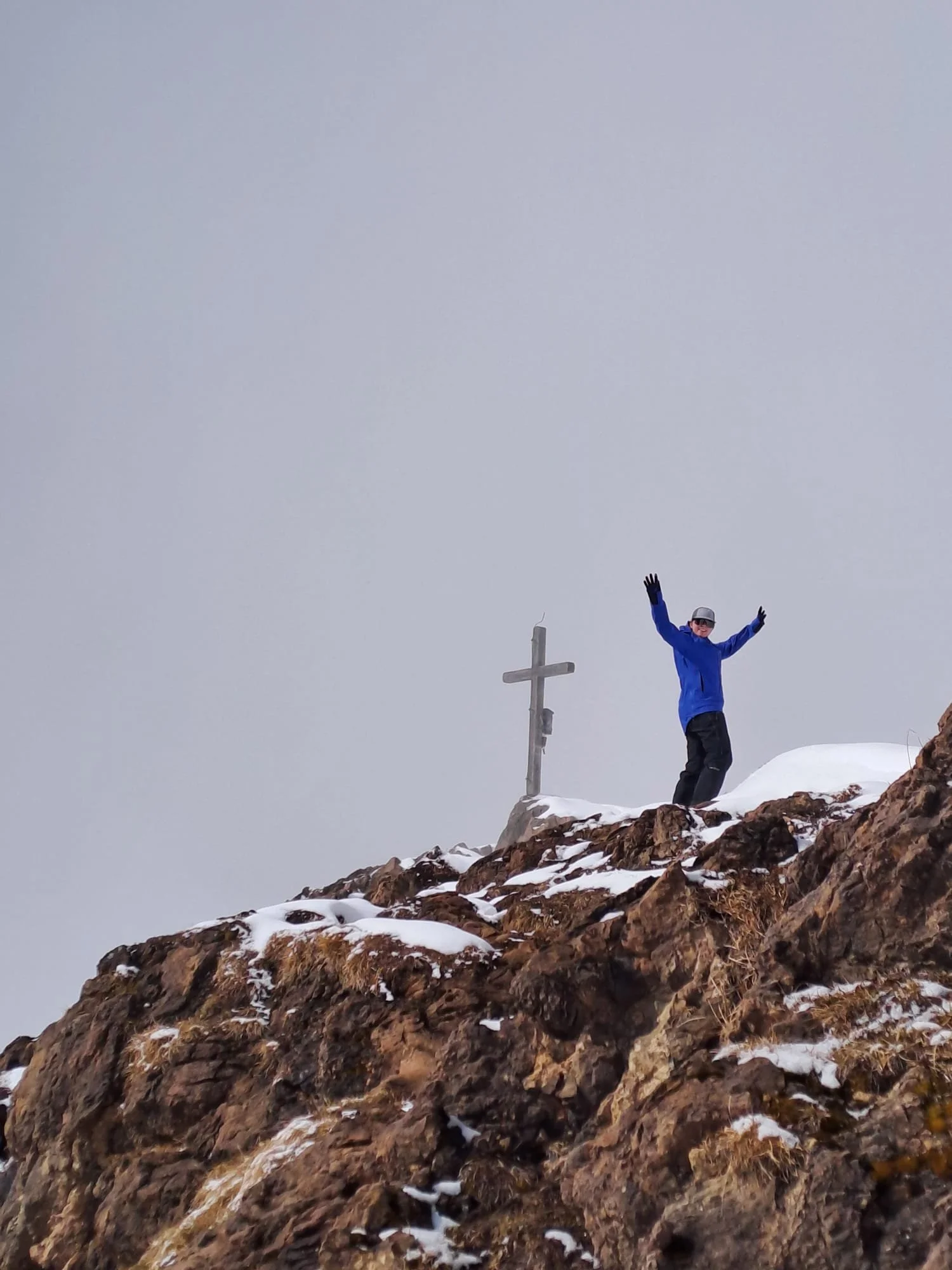 A person wearing a blue jacket, black pants, a helmet, and gloves standing on a snowy rocky mountain slope with a cross in the background and arms raised.