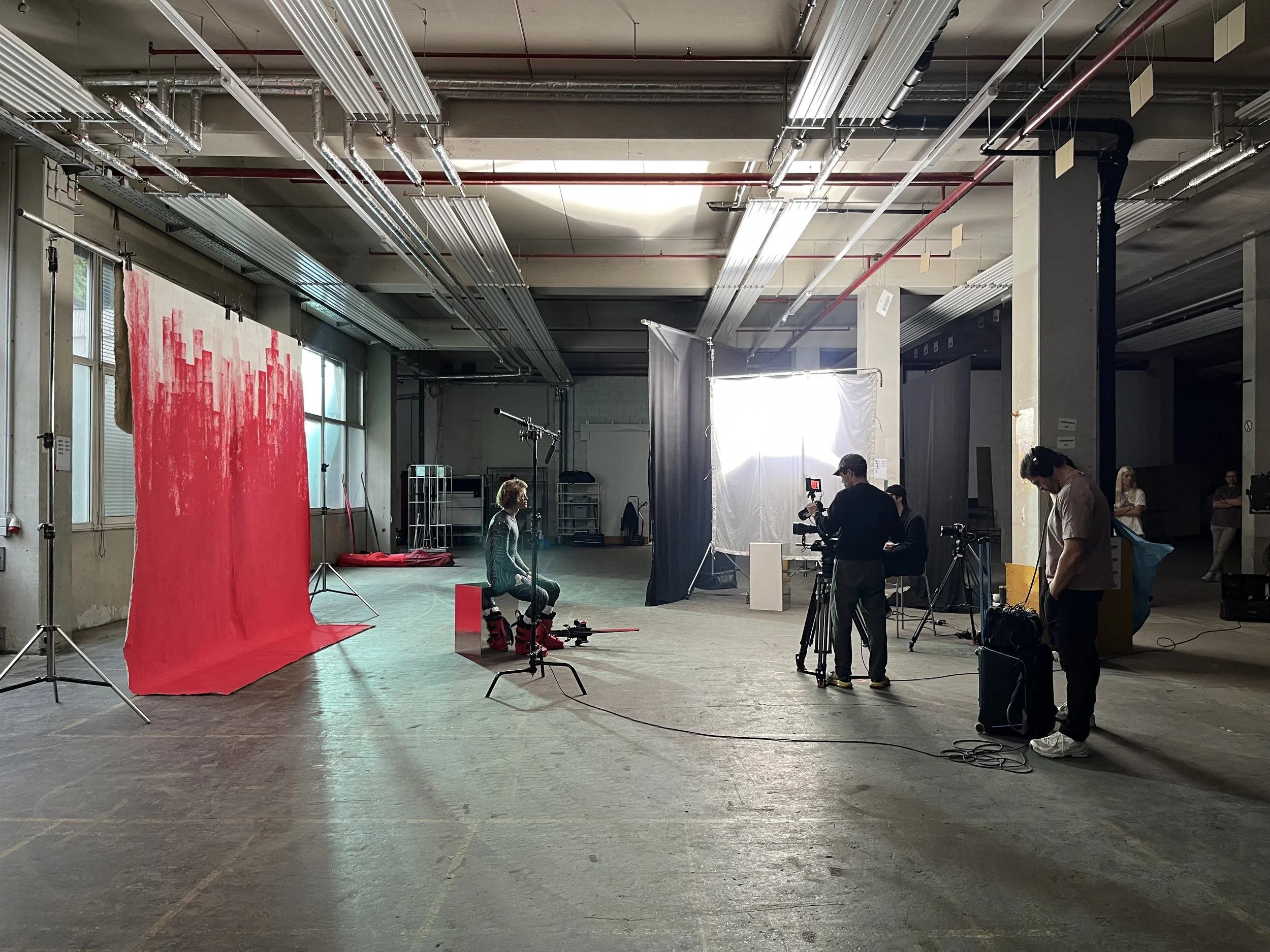 A photo shoot setup inside a large industrial-style space with concrete floors and ceiling. There is a red backdrop on the left side, and a woman is seated in front of it. Studio lighting and camera equipment are visible, including a crew member operating a camera and others working around the scene.