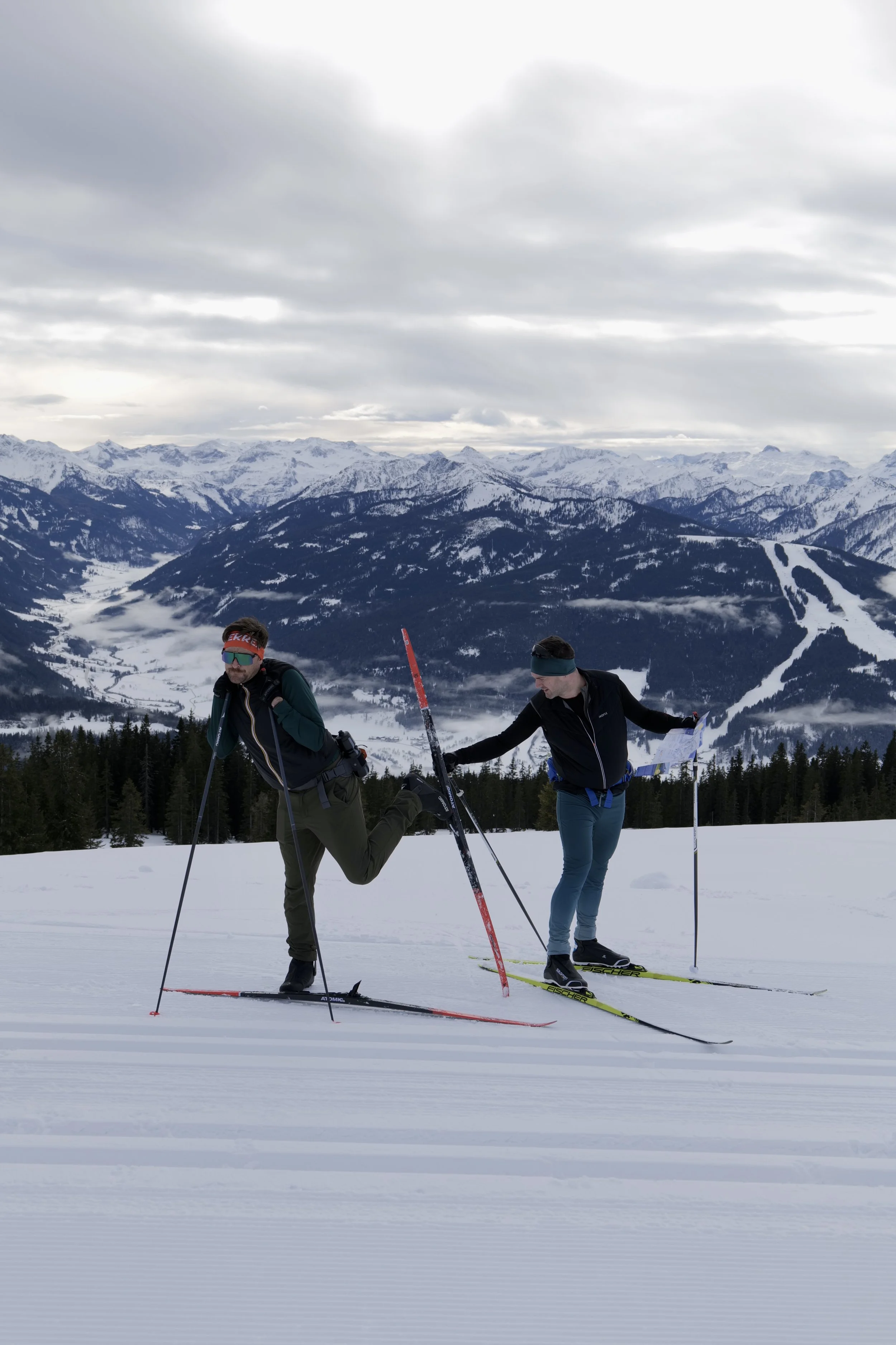 Two people cross-country skiing on a snow-covered trail with a mountain range in the background under cloudy skies.