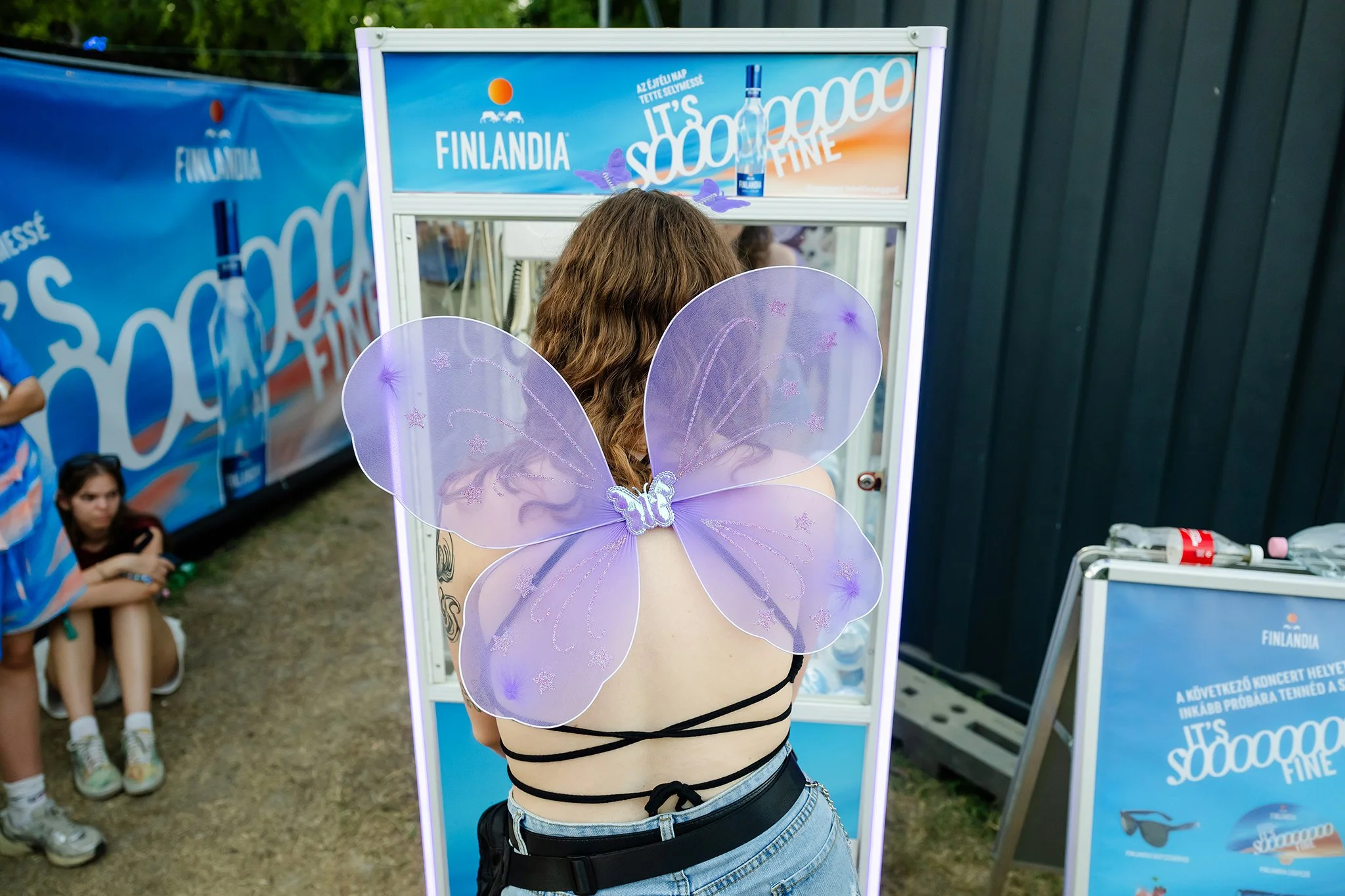 A woman wearing purple fairy wings and a black string top stands in front of a mirror at an outdoor event promoting Finlandia vodka, with other people sitting nearby and a blue advertising banner in the background.