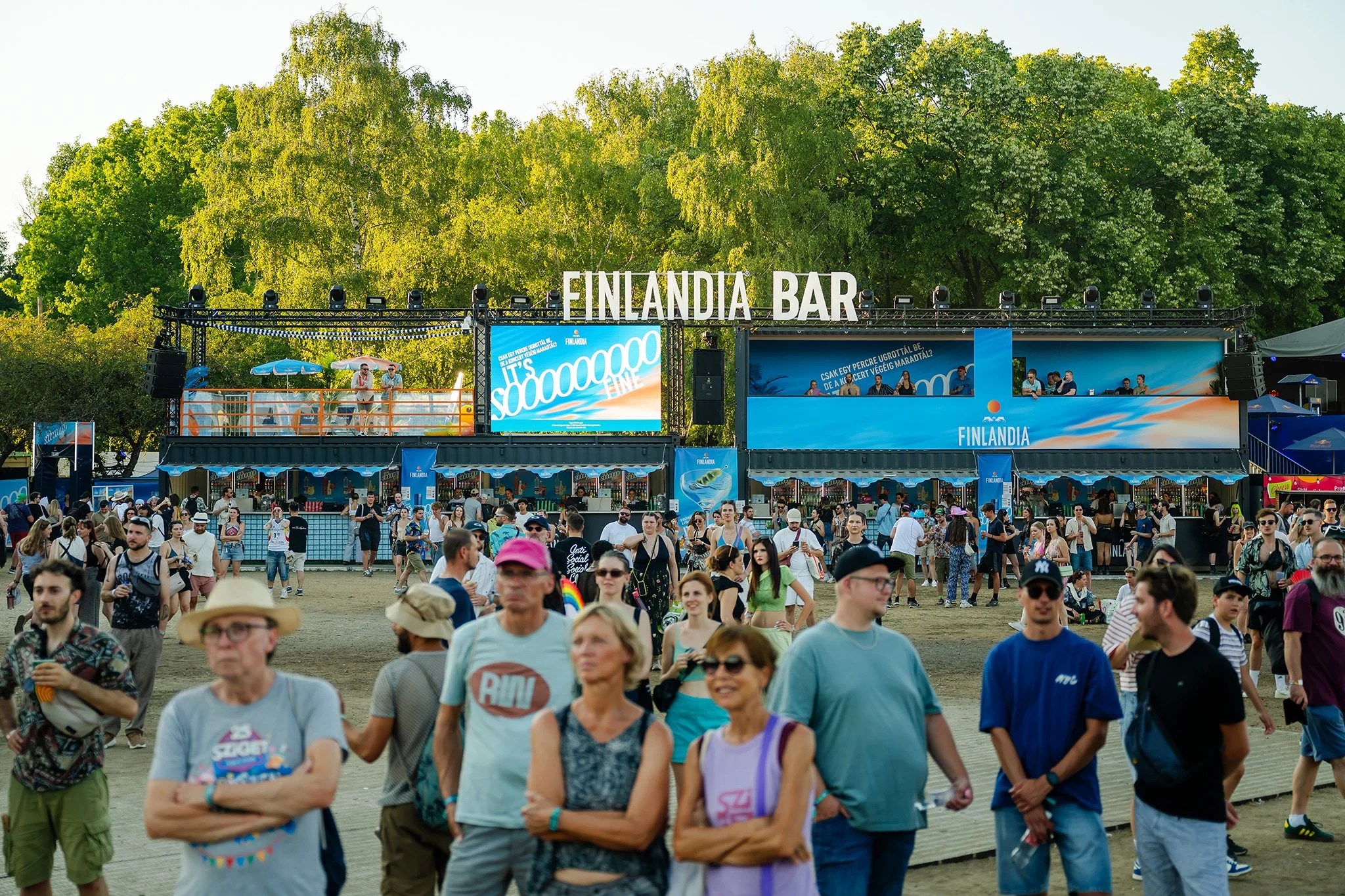 Crowd of people gathered at an outdoor festival with a stage in the background labeled 'Finlandia Bar' and large screens displaying promotional content, surrounded by green trees under a clear sky.