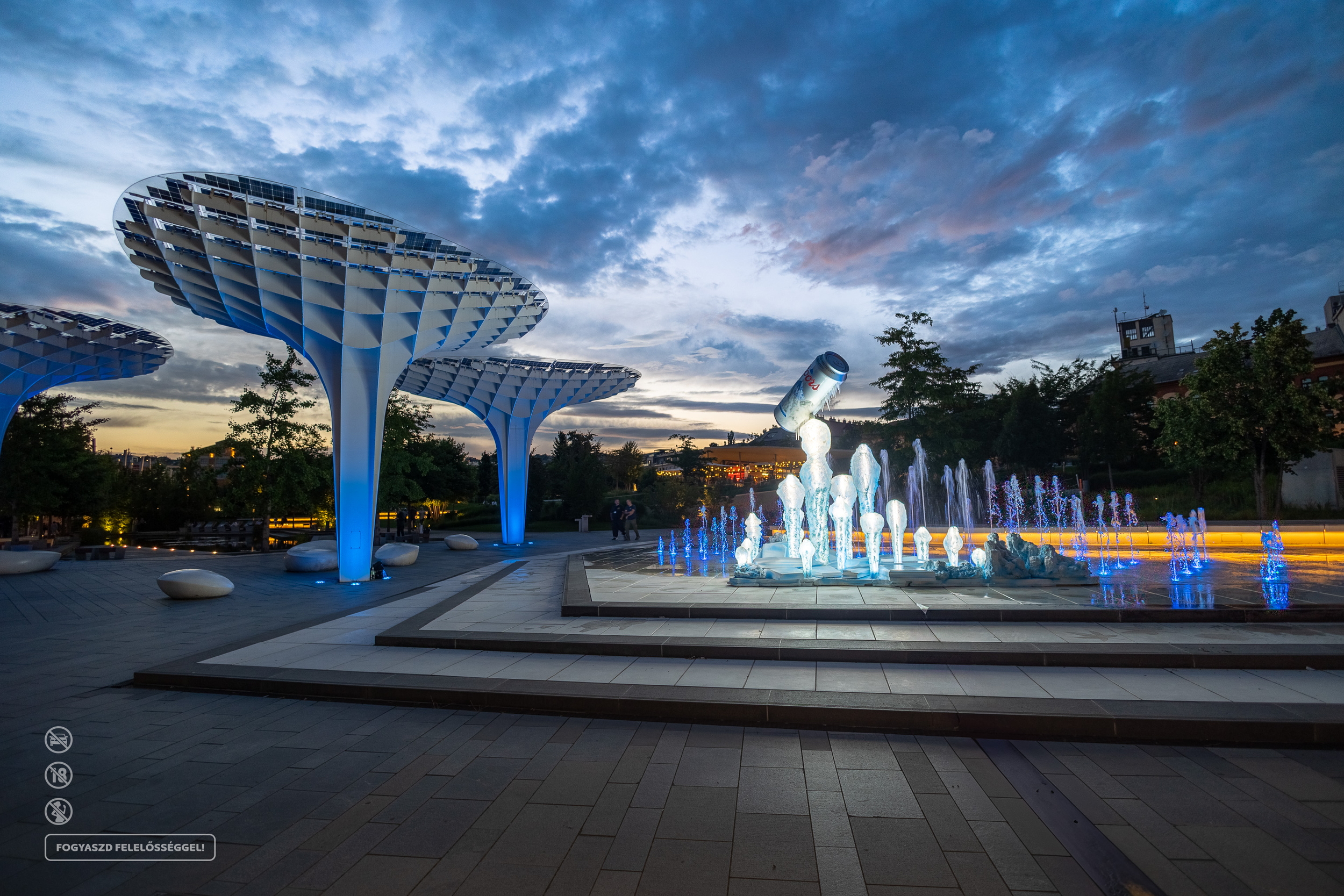 Ourdoor park with modern futuristic pavilion structures with multiple tiers, illuminated with blue lighting, located near a fountain with water jets, at dusk under a partly cloudy sky, trees, and city buildings in the background.
