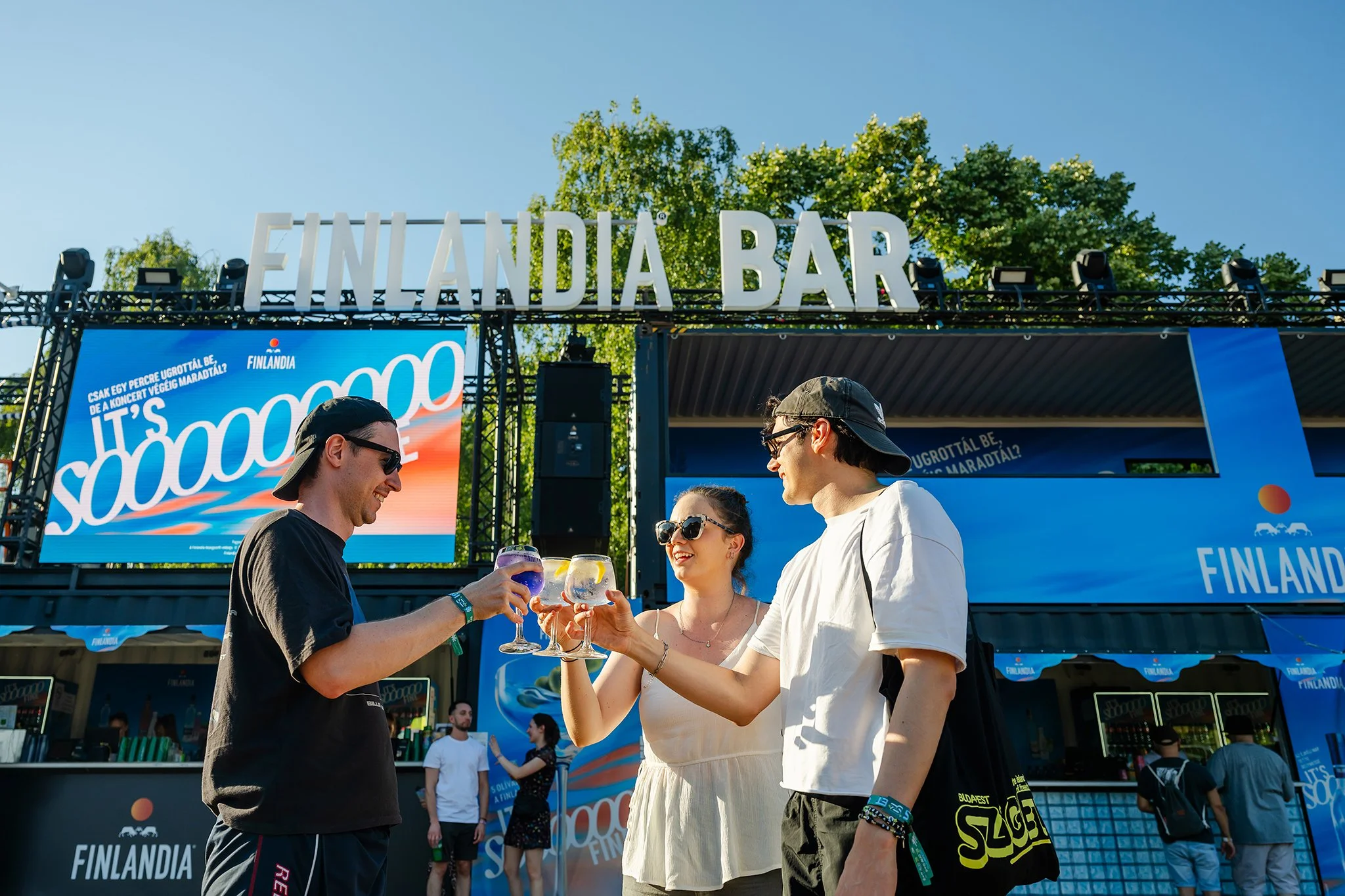 People at Finlandia event holding glasses of drinks outdoors, with a festival banner overhead and blue sky in the background.
