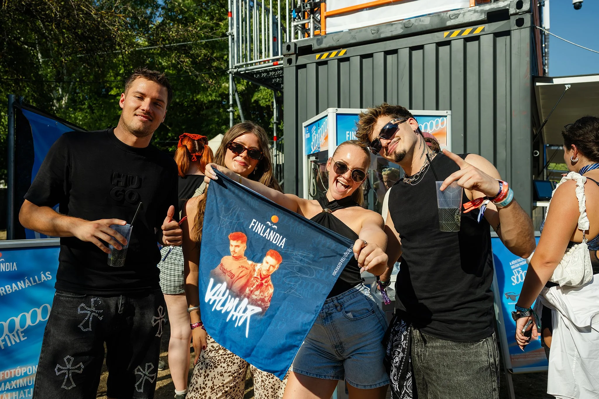Group of five young adults smiling and posing for a photo at an outdoor event, holding a blue banner with images of two men and text related to Finland, with trees and event structures in the background.