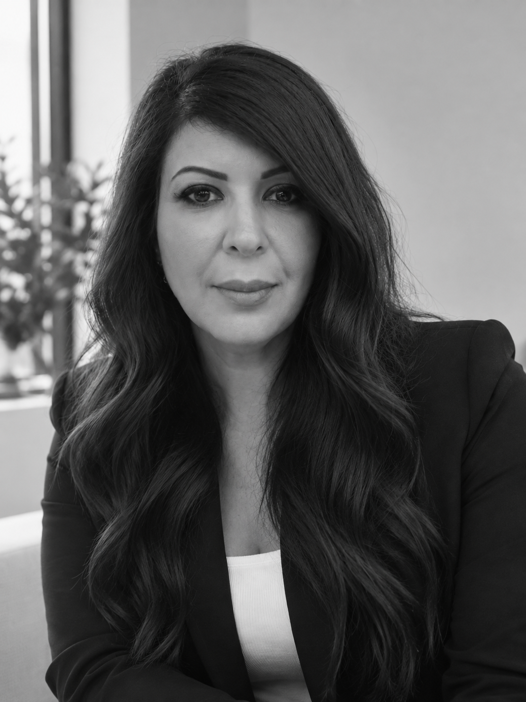 Black and white photo of Klara Yousif with long wavy dark hair, wearing a blazer, sitting indoors with a window and plant in the background.