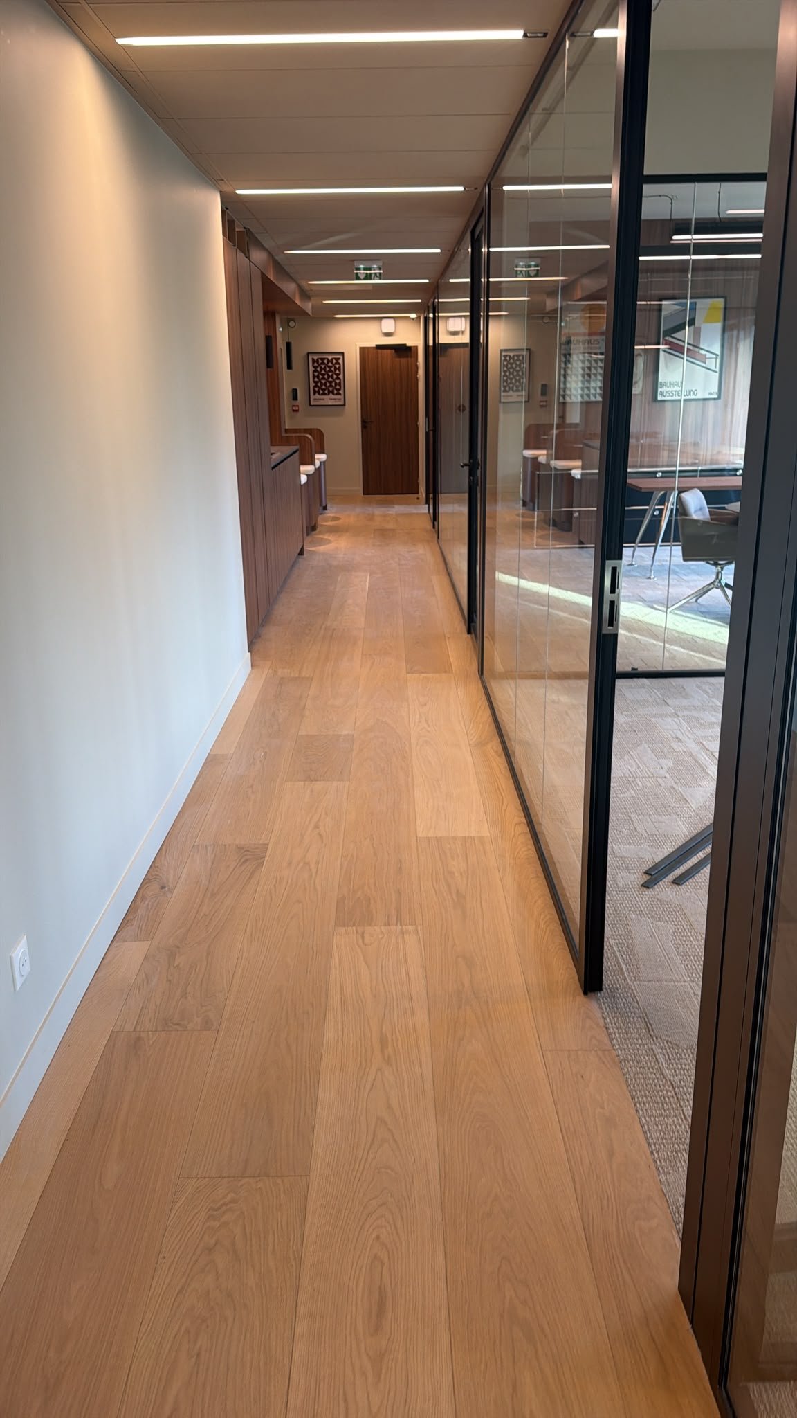 Couloir moderne de bureaux avec parquet en bois, cloisons vitrées et éclairage encastré.